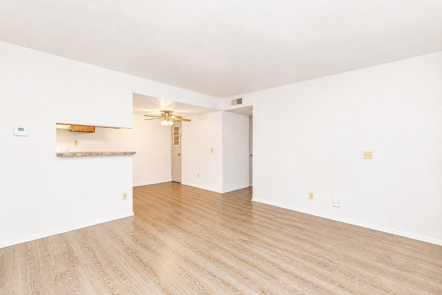 Empty living space with light-colored walls and wood-like flooring. A small kitchen area with a counter is visible on the left, and a ceiling fan is installed in the center of the room. The area is well-lit, creating a spacious and inviting atmosphere.