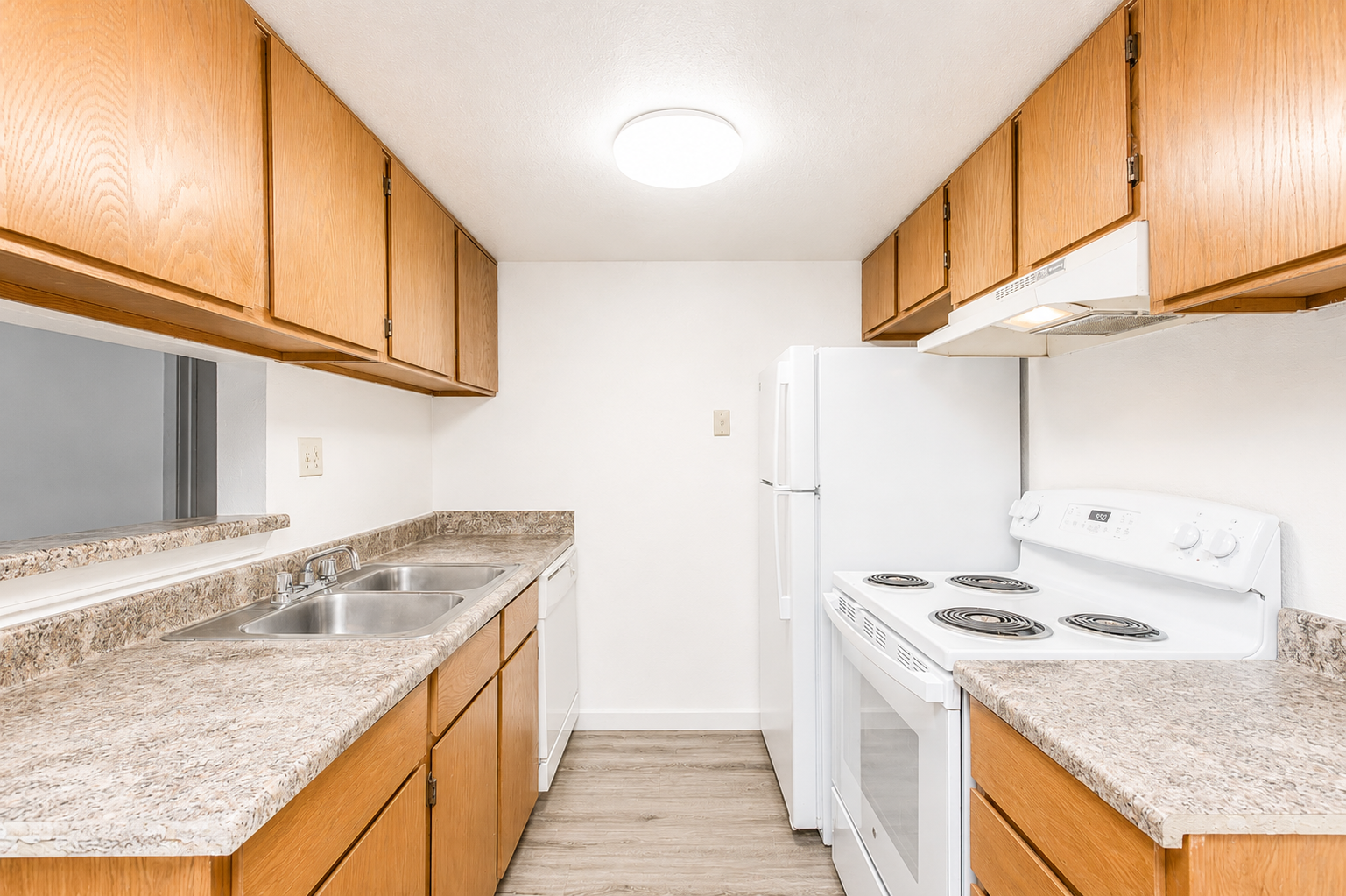Kitchen featuring light wood cabinetry, a double sink, and granite countertops. An electric stove and oven are situated next to a white refrigerator. The room has neutral-colored walls and a ceiling light, creating a bright and clean atmosphere.