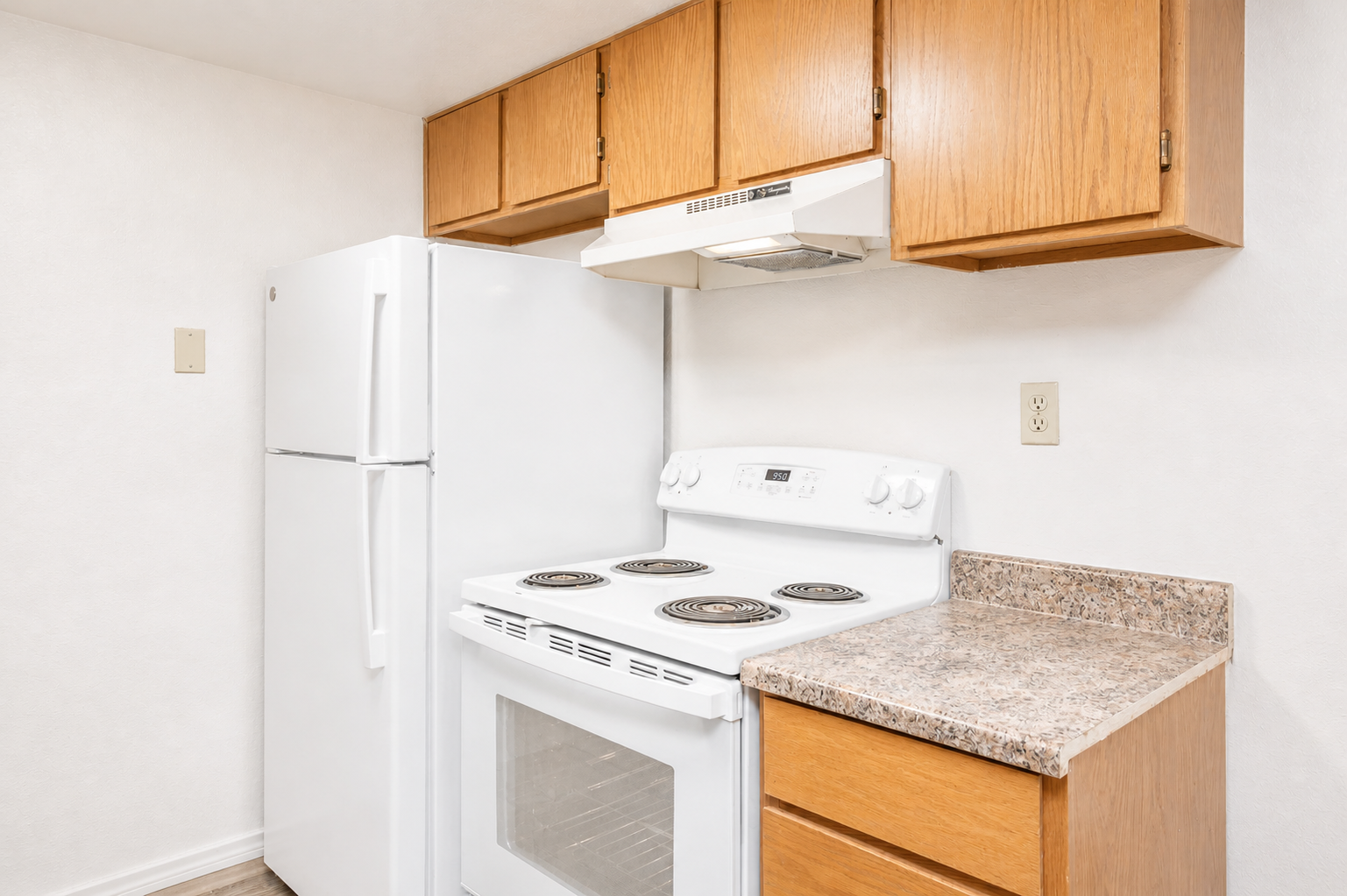 A compact kitchen featuring white appliances, including a refrigerator, stove, and exhaust hood. The cabinetry above the countertop has wooden doors, and the countertop is made of a speckled stone material. The overall color scheme is light and neutral, creating an inviting atmosphere.