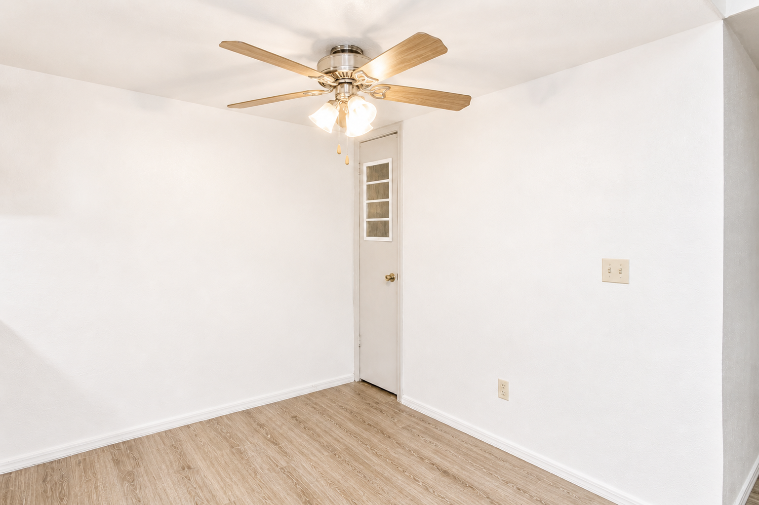 A spacious room with light-colored walls and a wooden floor. The ceiling features a ceiling fan with lights, and there is a white door with glass panels leading to another area. The room is empty, providing a clean and simple aesthetic.