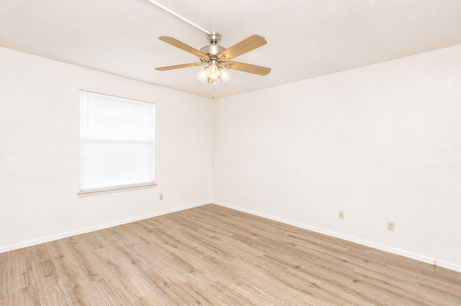 Empty room with light-colored walls and wooden flooring. It features a ceiling fan with wooden blades and a single window with closed blinds, allowing soft natural light to enter. The space is well-lit and uncluttered, creating a blank canvas for decoration.