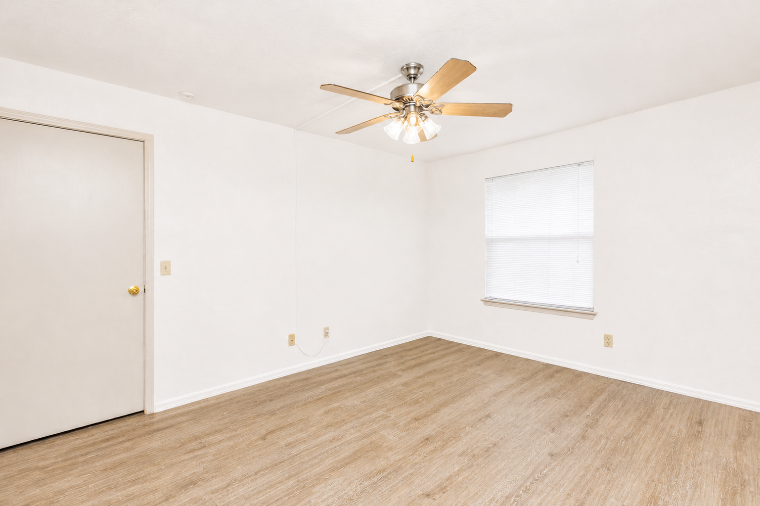 A bright and empty indoor room featuring light-colored walls and laminate flooring. The ceiling has a ceiling fan with wooden blades. A single window with blinds allows natural light to enter, and there is a closed door leading to another space. The room is uncluttered and ready for furnishing.