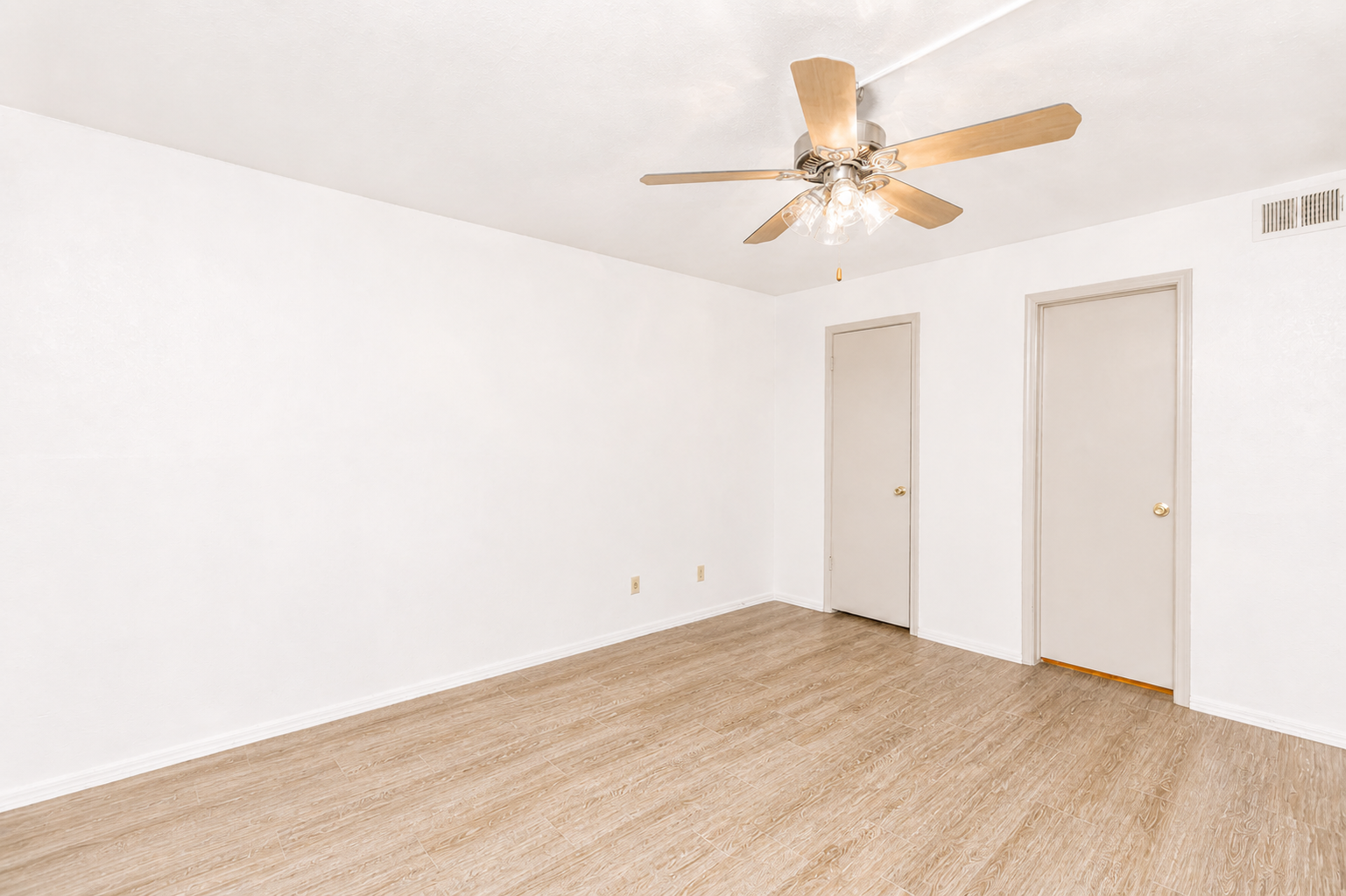 A bright, empty room with light beige walls and a ceiling fan. The floor is a light wood laminate. There are two closed doors on the right side, and the space has a clean, minimalist appearance, suitable for various purposes like a living area or office.
