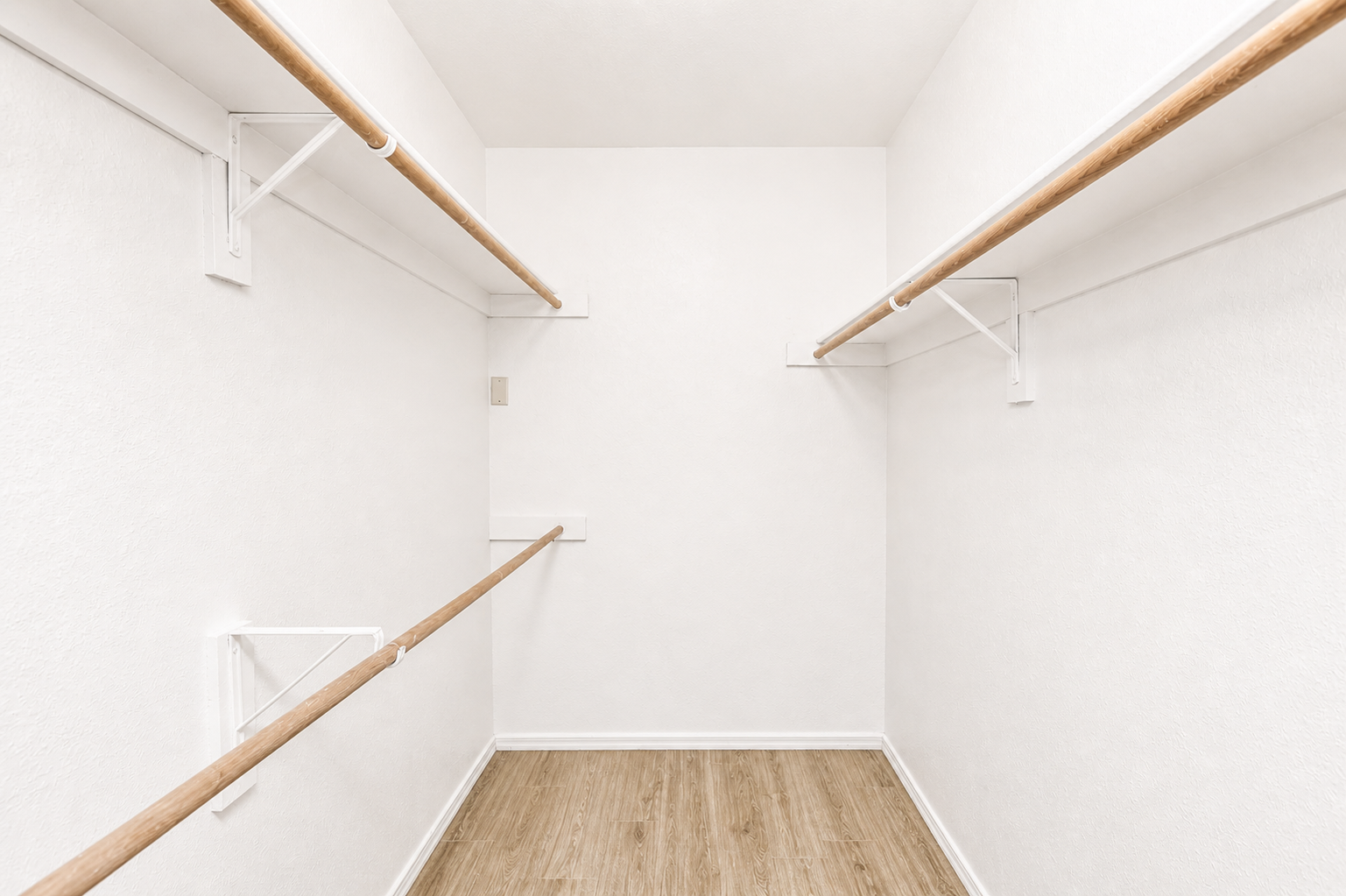 A spacious, empty walk-in closet featuring two wooden rods for hanging clothes, white walls, and a light wooden floor. The lack of shelves or additional storage makes the space feel open and minimalistic.