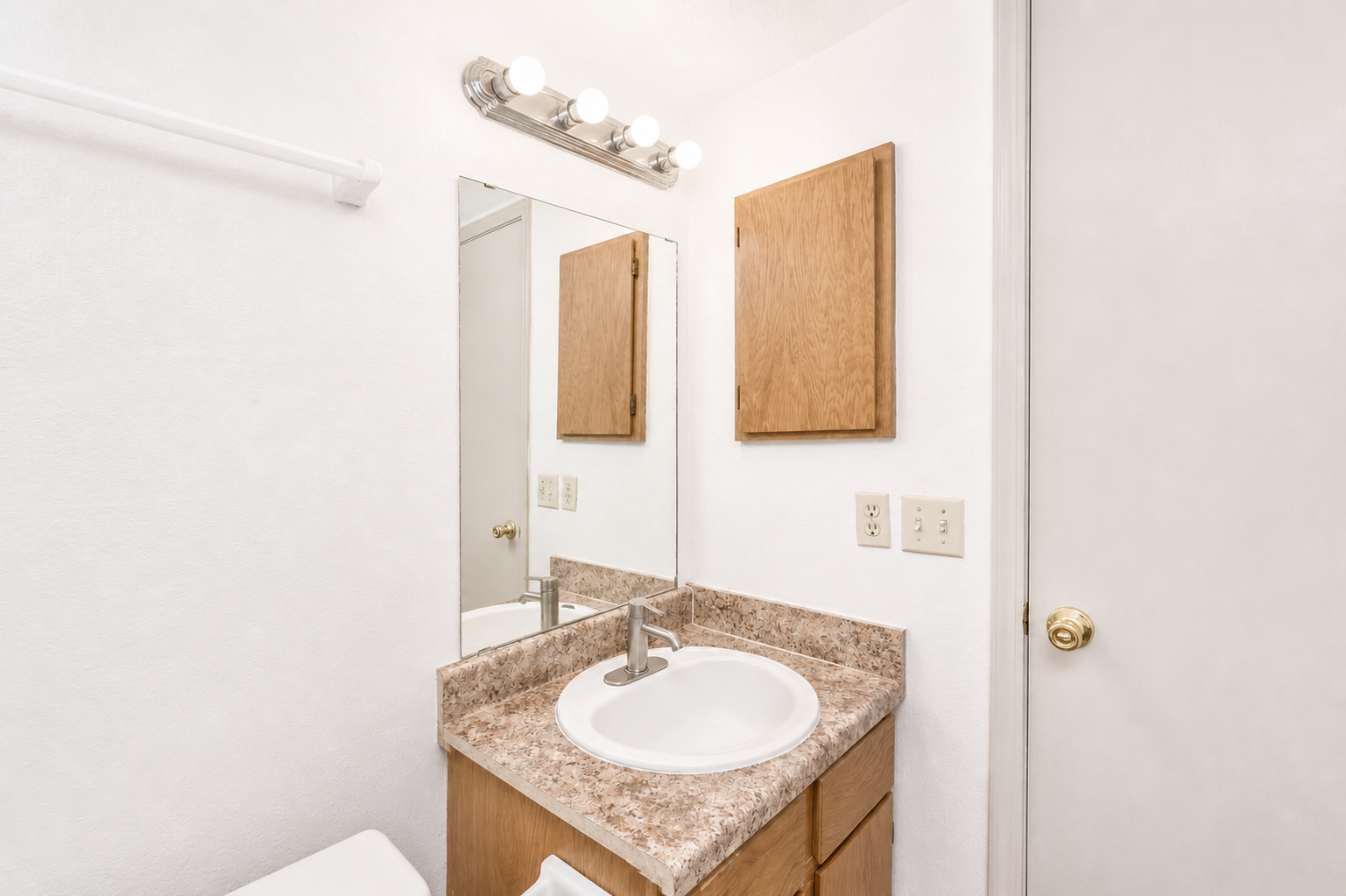 A small bathroom featuring a beige countertop with a circular sink, a mirror above it, and wooden cabinets on the wall. Light fixtures are installed above the mirror, and the walls are painted white. A towel rack is visible on the left, and the door is closed to the right.