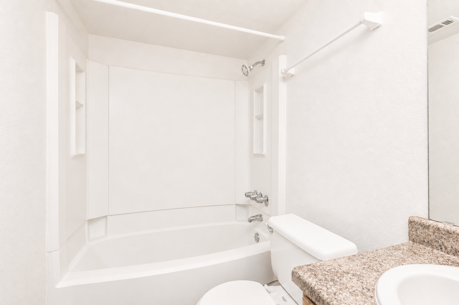 A clean, modern bathroom featuring a bathtub with a shower, white walls, and a granite countertop. The space includes a toilet and shelves for storage, creating a bright and minimalist design.