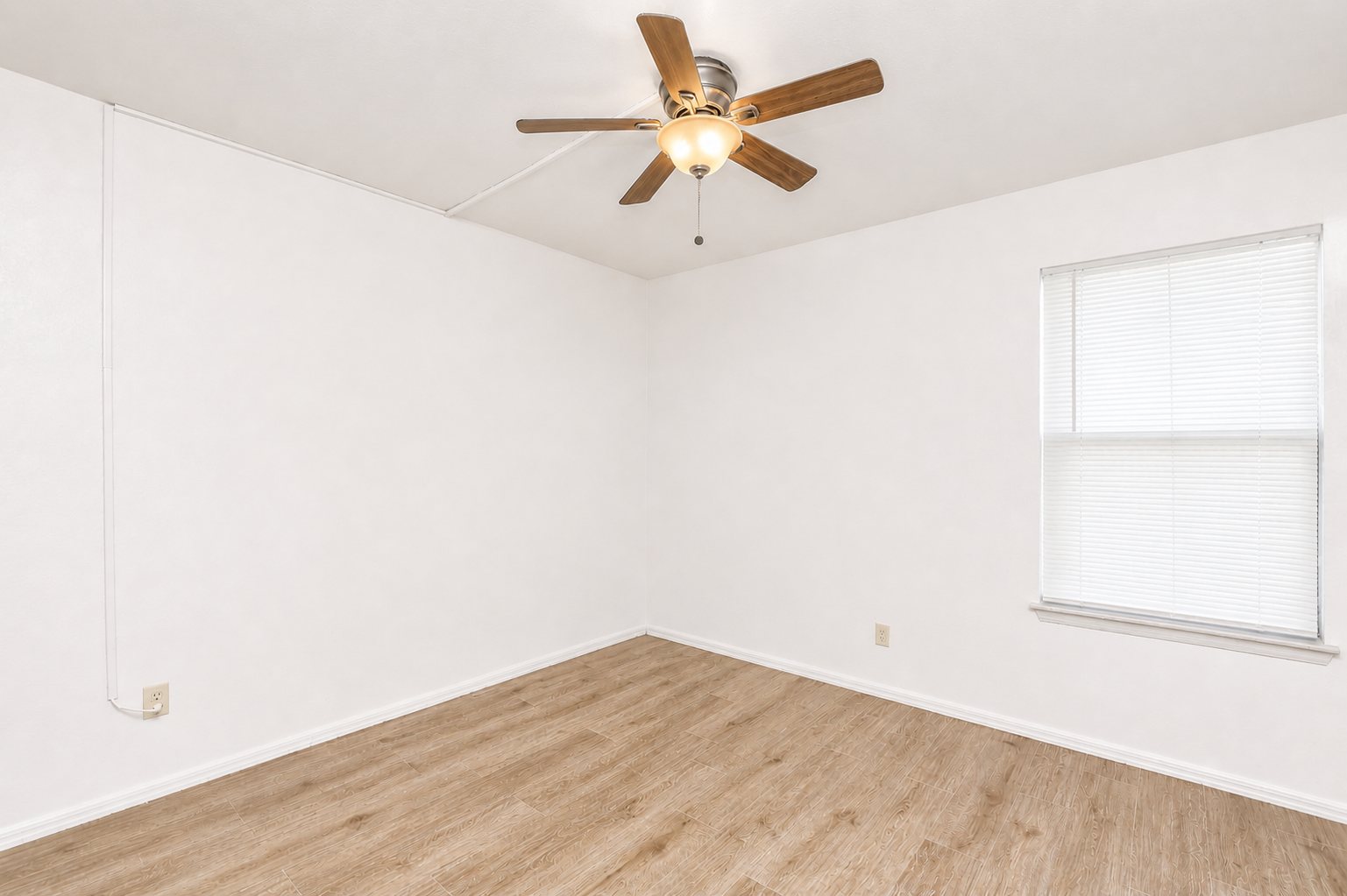 A spacious, empty room featuring light-colored walls, a ceiling fan with wooden blades, and a window with blinds. The floor is a light wood laminate, and there are no furnishings present, creating a clean and minimalistic space.