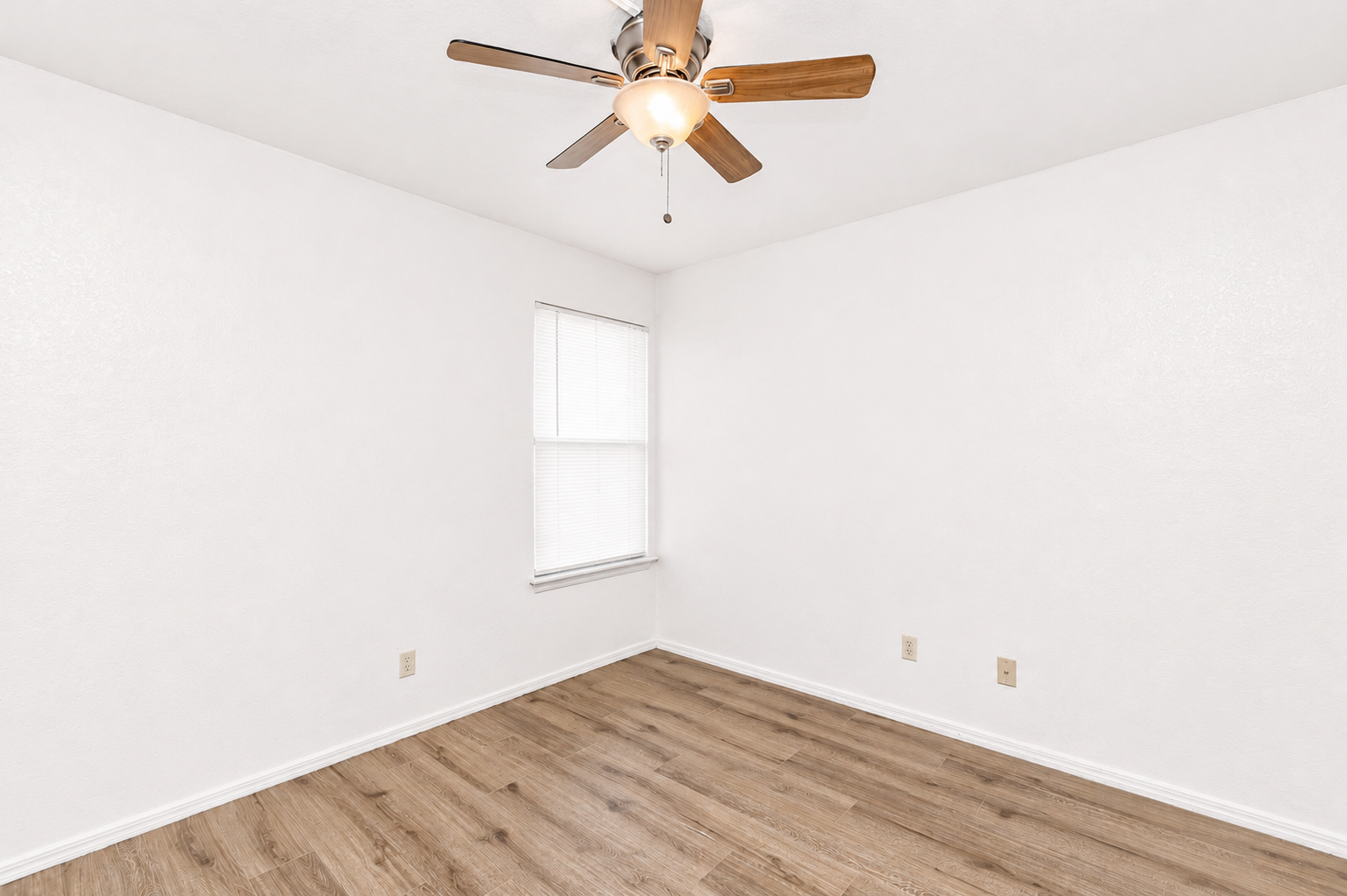 Empty room with light-colored walls and a wooden floor. A ceiling fan with wooden blades is visible. There is a window with blinds on one wall, allowing natural light to enter. The overall space is simple and uncluttered, creating a bright and airy feel.