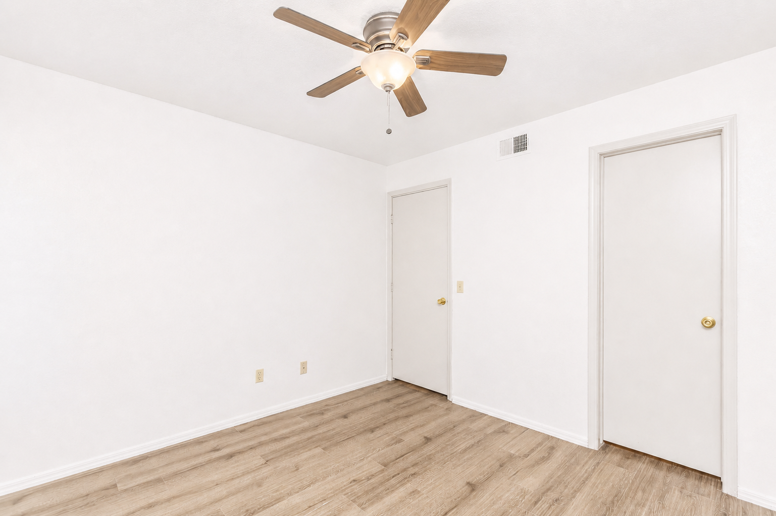 Empty room with white walls and a ceiling fan. The floor is made of light-colored wood. There are two doors on the walls, one visible closed door and another partially open. The space is bright and has a clean, minimalist look, suitable for various uses.