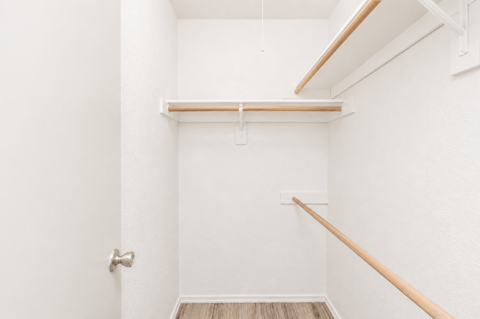 A clean, empty closet with white walls and wooden hanging rods. The floor is light-colored laminate. There is a closed door with a silver doorknob on the left, and a ceiling light fixture is present. The space is well-lit and tidy, suggesting it is ready for storage or organization.