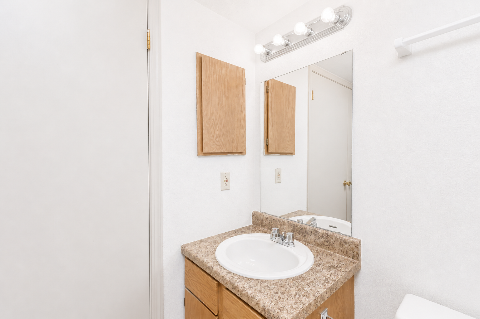 A clean, modern bathroom featuring a circular sink on a countertop with a speckled design. There are two wooden cabinets above the sink, a large mirror, and a light fixture with multiple bulbs. The walls are painted in a light color, creating a bright and spacious appearance.