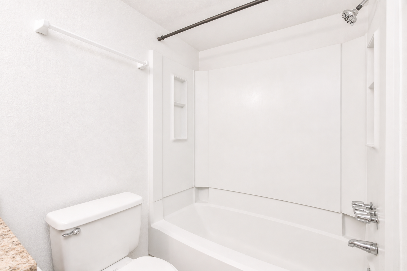 A clean, modern bathroom with white walls and fixtures. It features a bathtub with a shower head, a toilet, and a small shelf area in the wall. The overall design is minimalist and bright, creating an inviting space.