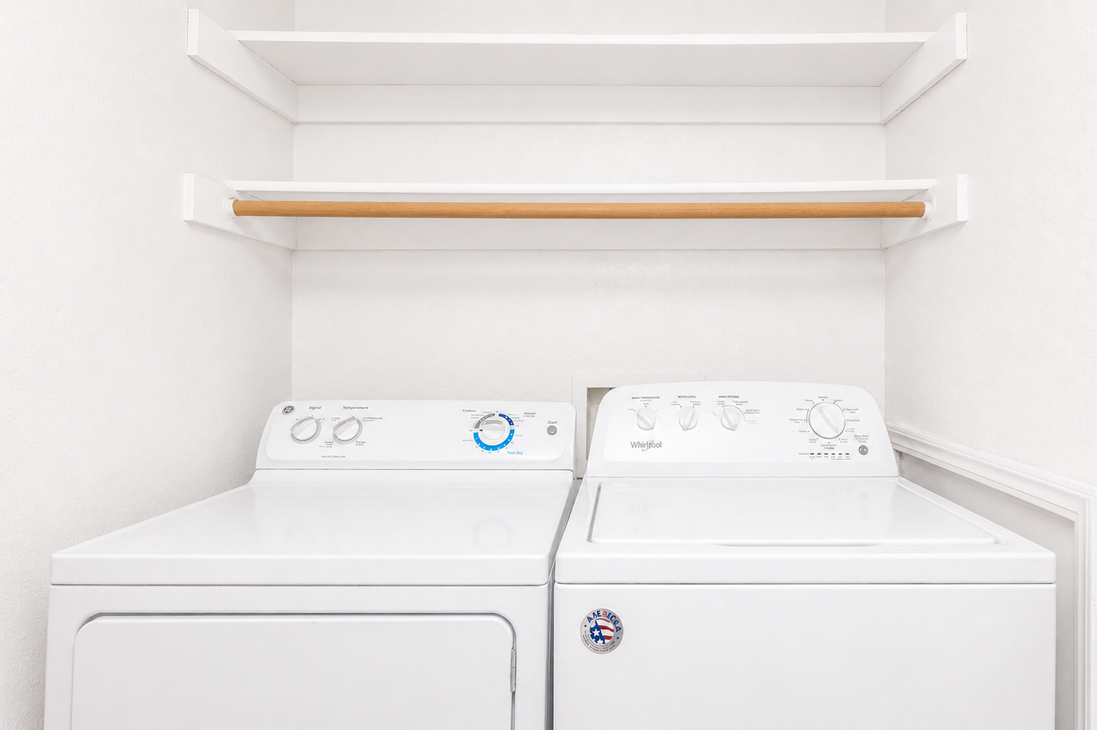A clean laundry room featuring a white washing machine and dryer side by side, with control panels visible. Above them, there is a simple wooden shelf mounted on the wall, providing storage space. The walls are painted in a light color, contributing to a bright and tidy atmosphere.
