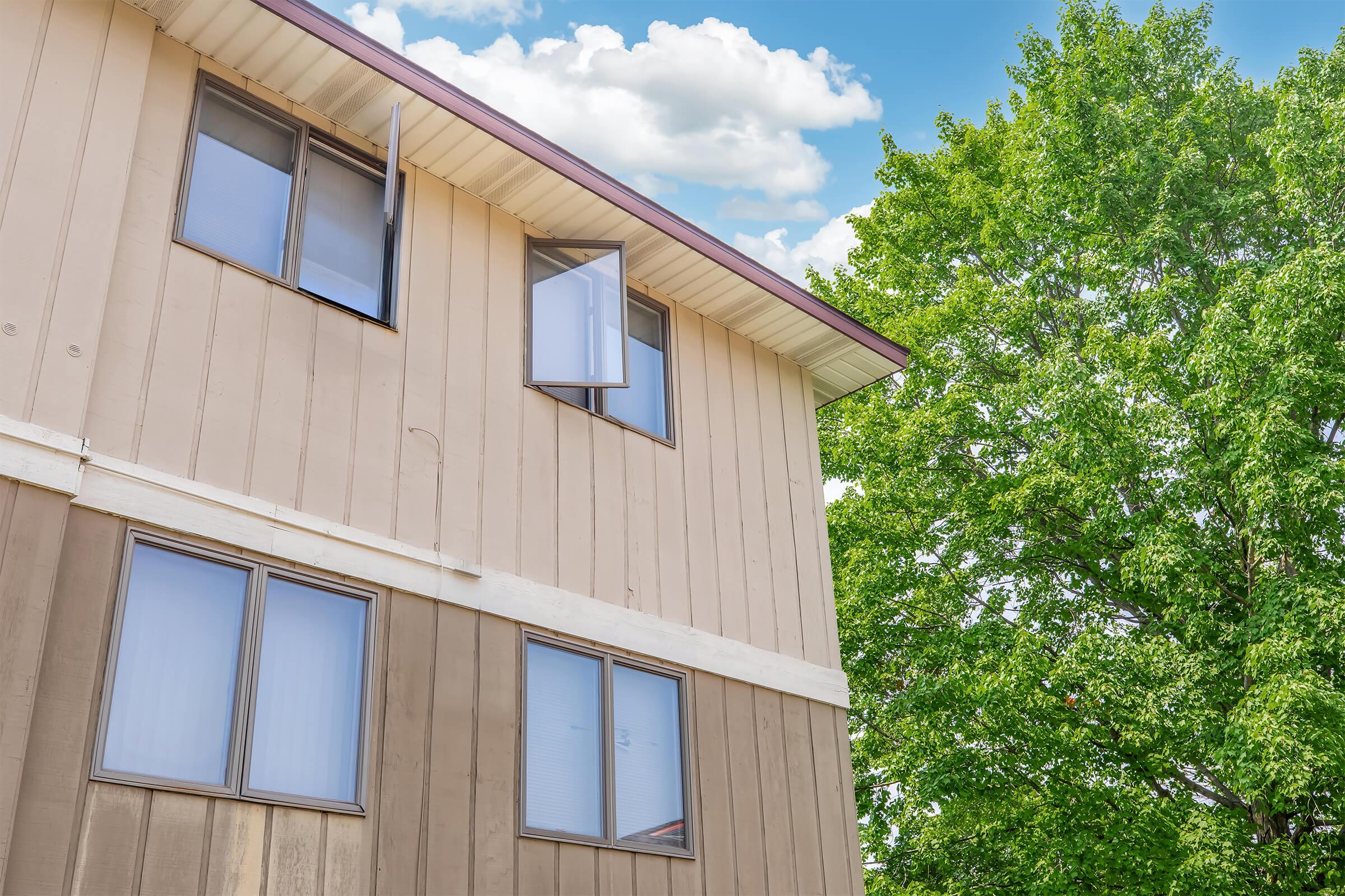 A two-story building with a wooden exterior and several windows. One window is open, allowing a view of the blue sky with scattered clouds. To the right, there is a large green tree, providing shade and contrast to the building's warm colors. The scene suggests a pleasant, sunny day.