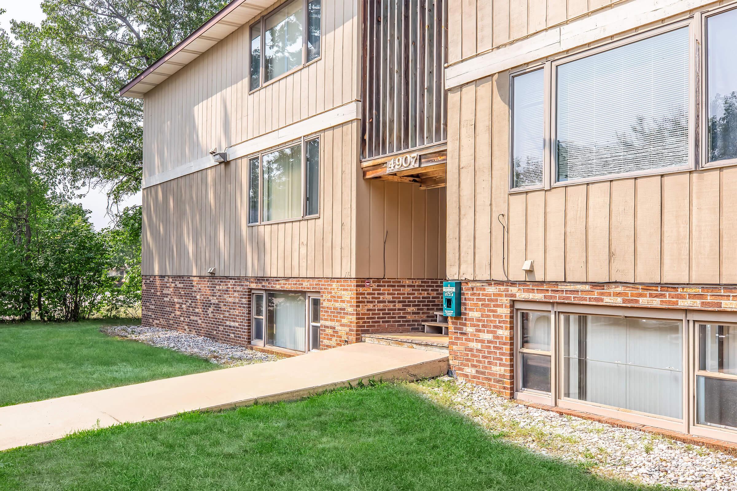 Exterior view of a two-story apartment building featuring a combination of siding and brick, with a green lawn in front. A walkway leads to the entrance, which has a wooden overhang and a mailbox mounted nearby. Surrounding trees provide a natural backdrop. Windows are large, allowing for ample sunlight.