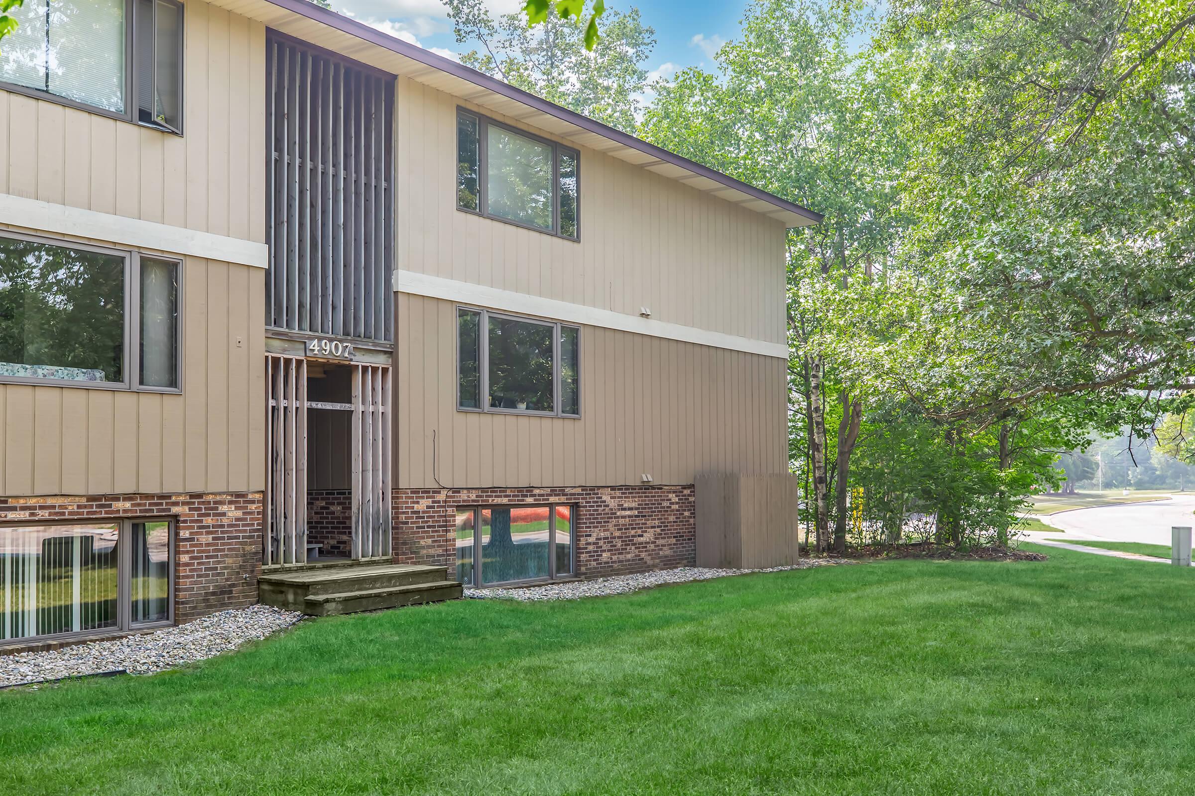 Exterior view of a modern two-story apartment building with tan siding and a brick base. The entrance features steps leading up to a patio area, surrounded by greenery and a well-maintained lawn. Trees are present nearby, and the setting appears sunny and inviting.