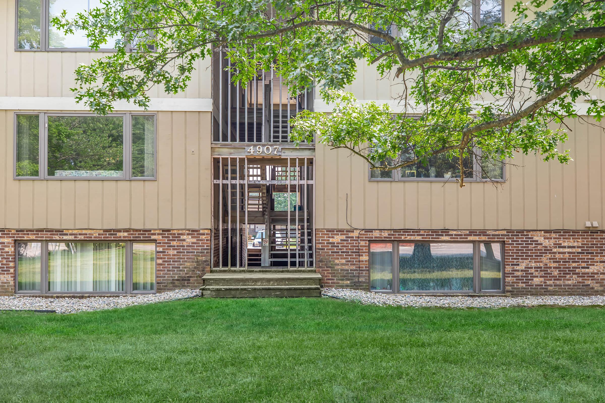 A two-story apartment building with a brown and beige exterior. The entrance features metal bars and a small staircase leading to it. Nearby, there's a large green lawn and several windows offering a view of the exterior landscape. There are trees and shrubs in the vicinity, enhancing the outdoor environment.