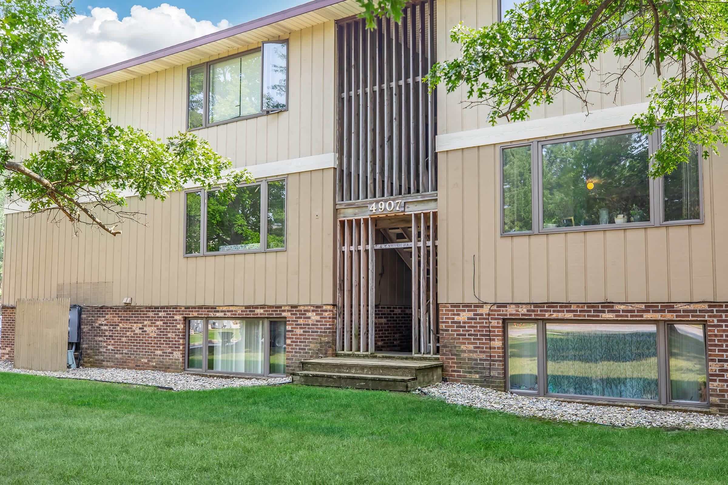 A two-story residential building featuring a mix of beige siding and brick. The entrance has a wooden deck with a slatted railing. Large windows on the front provide natural light, surrounded by well-maintained green grass and landscaping. Tree branches frame the image, adding a touch of greenery.