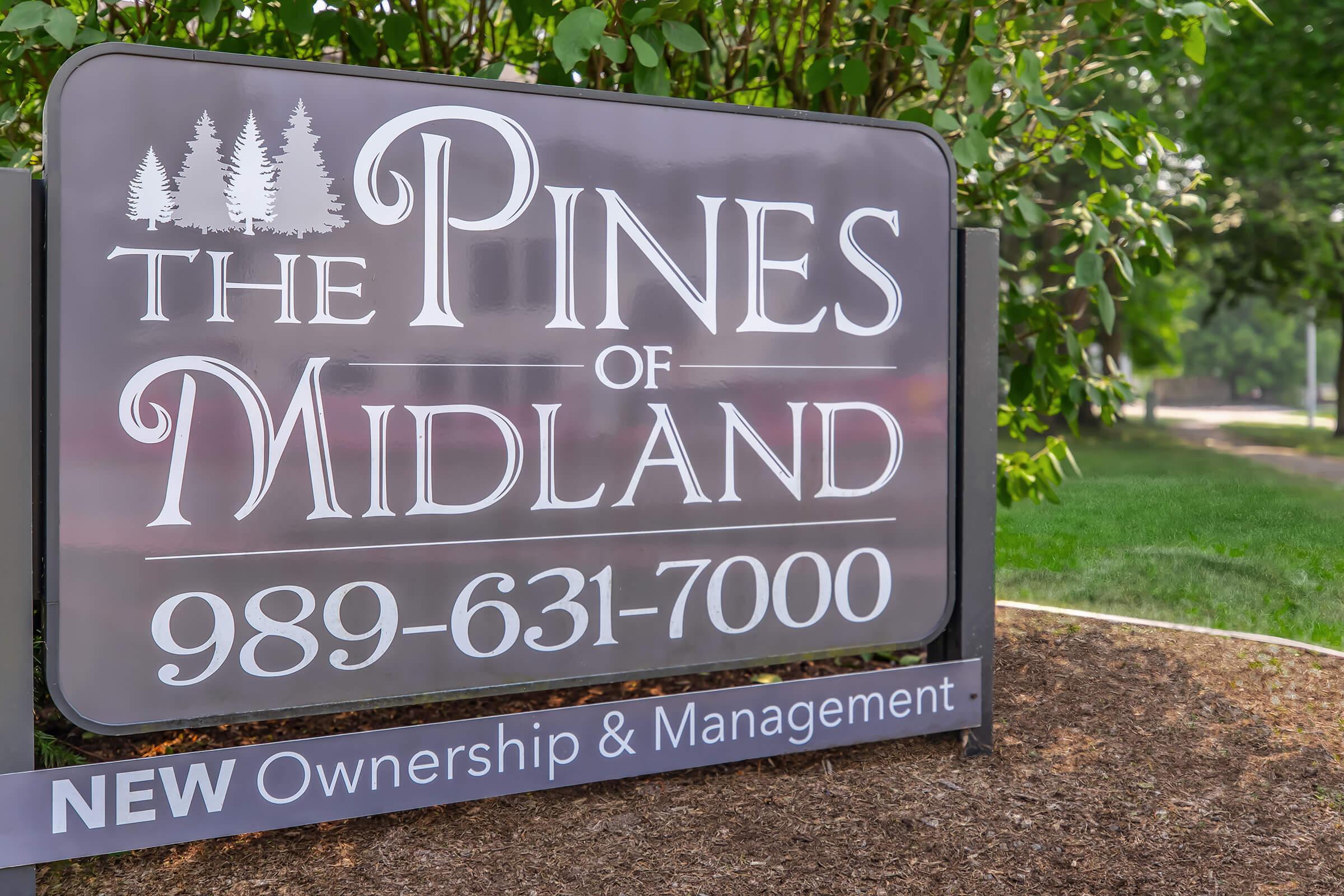 Sign for "The Pines of Midland" featuring the text clearly. Includes a phone number, "989-631-7000," and the phrase "NEW Ownership & Management." The sign is set against a green, landscaped background.