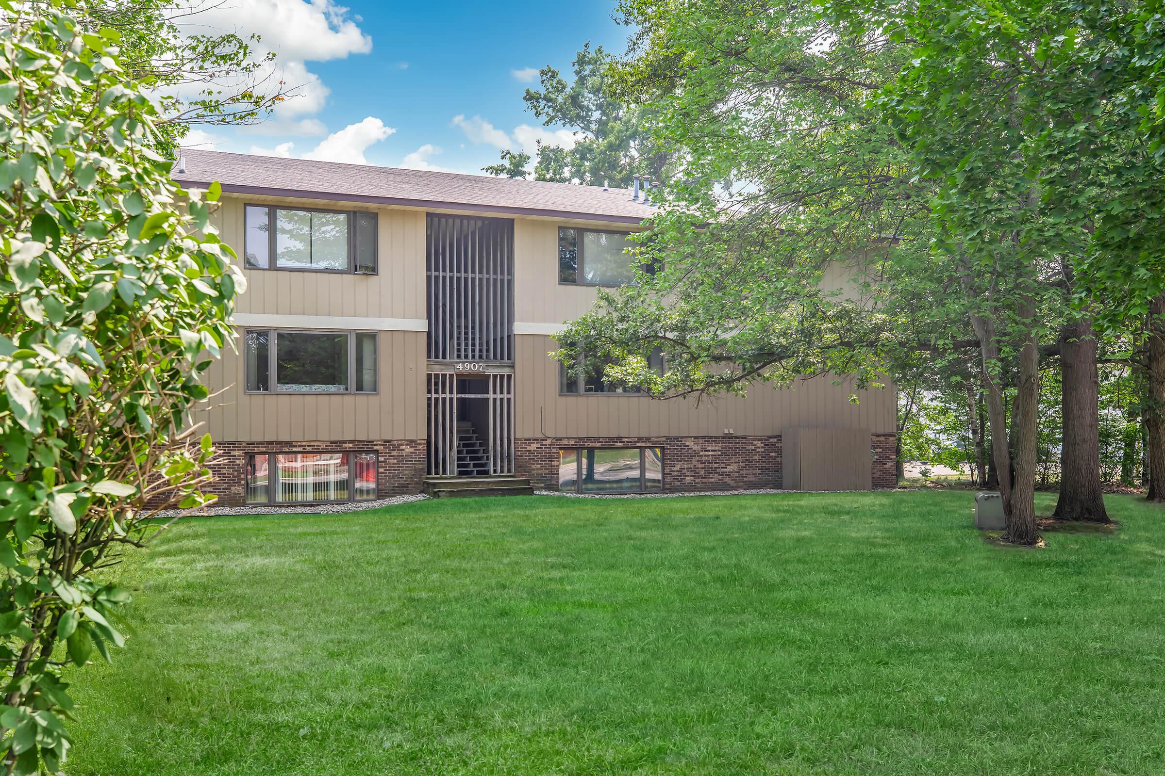 A modern two-story house with a beige exterior, featuring large windows and a brick foundation. The front yard is lush and green, surrounded by trees. A staircase leads to the entrance, and the sky is clear with a few clouds. The setting appears calm and inviting.