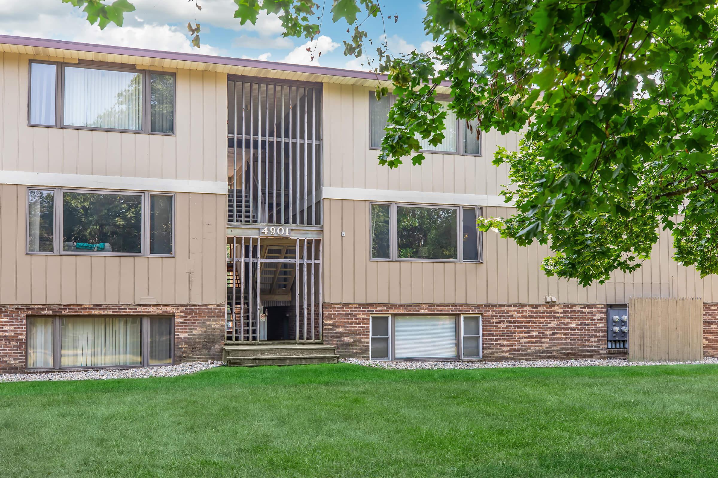 A multi-unit residential building with a beige and brown exterior. The structure features several large windows and a central entrance with metal railings. A well-maintained lawn surrounds the building, and trees provide some shade in the front.