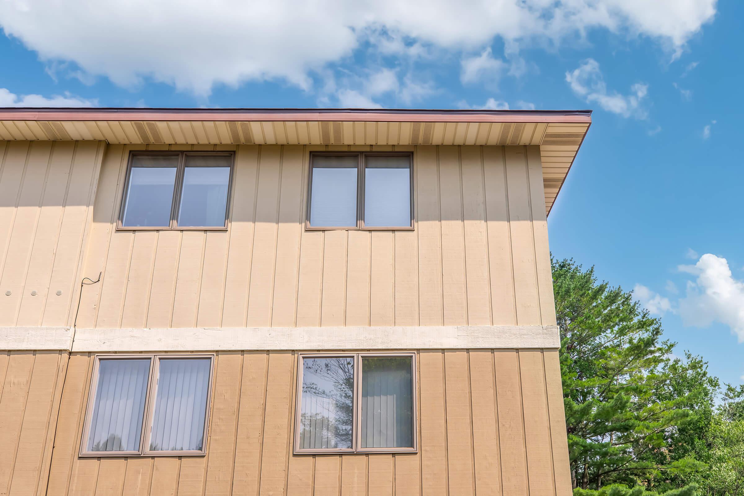 Two-story building with beige horizontal siding and a flat roof. The upper floor features two large, rectangular windows with dark frames. The background includes a clear blue sky with a few clouds and green trees at the bottom, creating a serene suburban atmosphere.