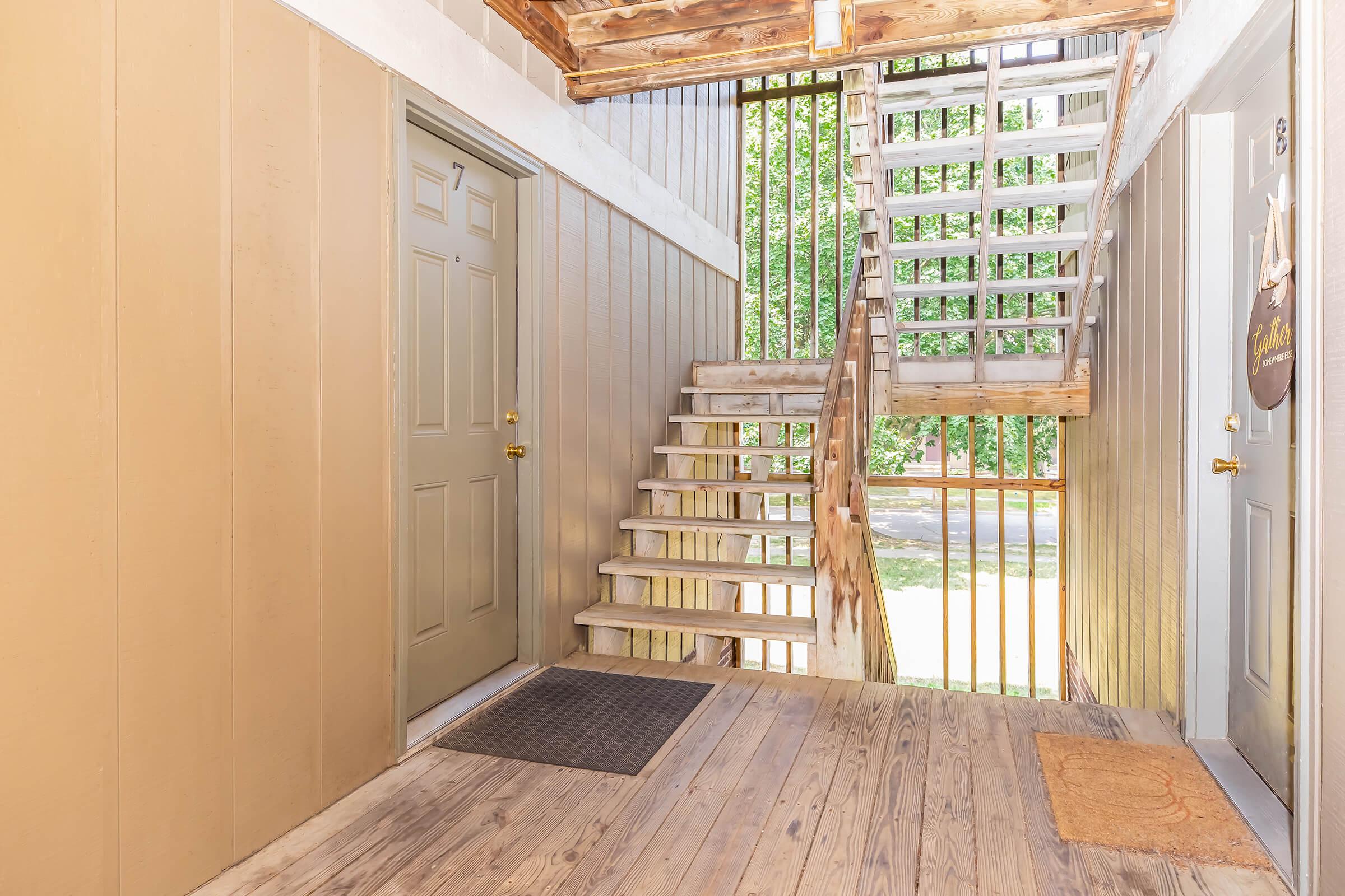 Interior view of an apartment building corridor featuring two doors on the left and a wooden staircase on the right leading to an upper level. Natural light floods the space, highlighting the wooden flooring and outdoor greenery visible through the open stairwell.