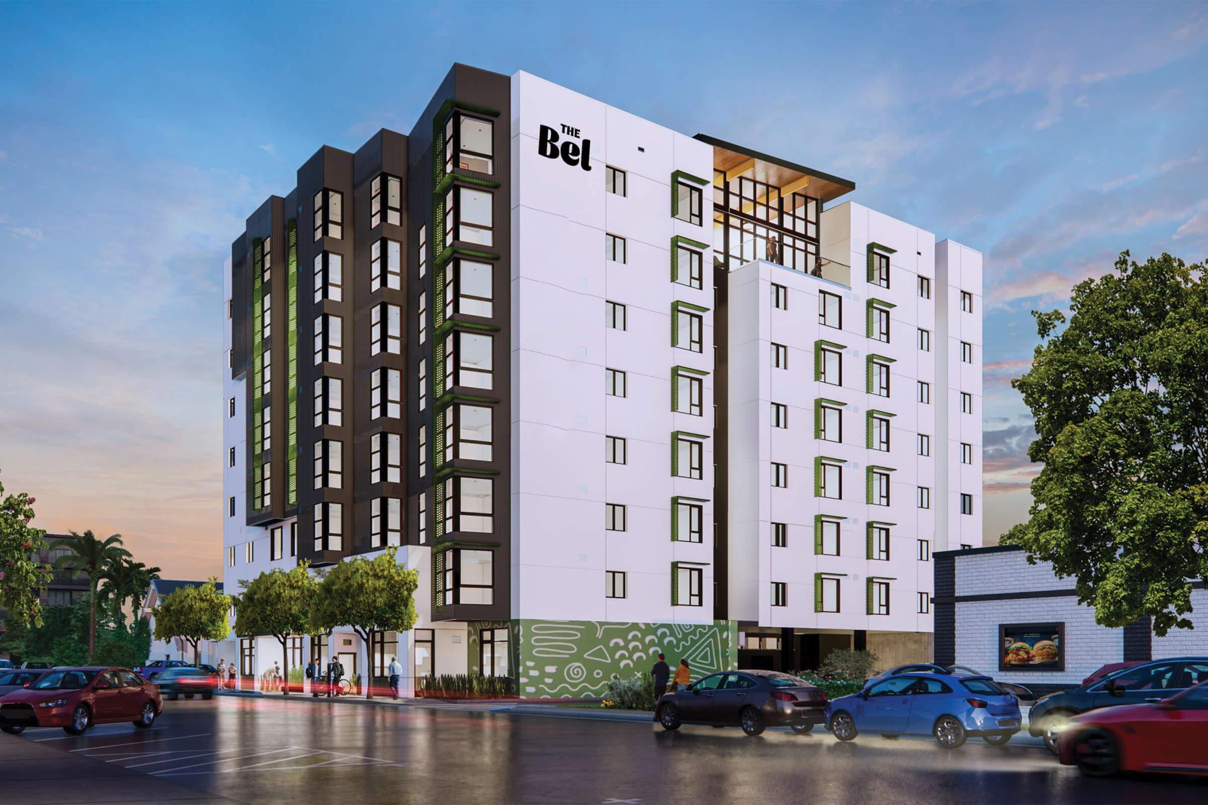 A modern multi-story hotel building, featuring a combination of white and dark gray panels. The structure has large windows and a contemporary design, with greenery around the entrance and cars parked in the foreground. The sky is partially cloudy, suggesting either dawn or dusk lighting.