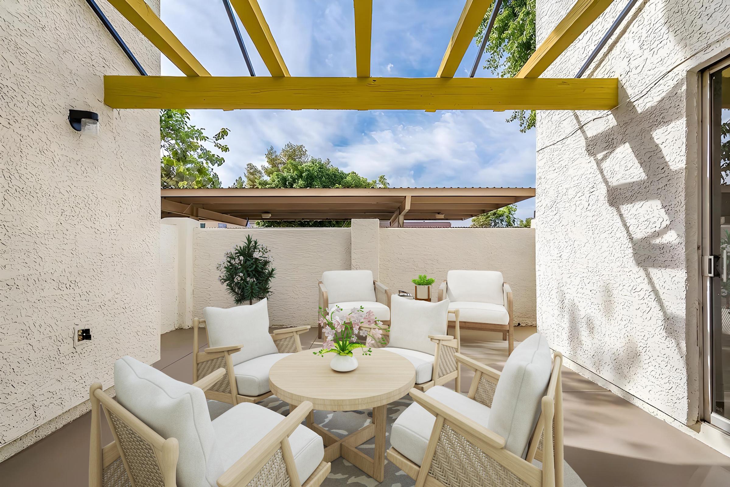A cozy outdoor patio area featuring a round wooden table surrounded by white cushioned chairs. Above, a yellow pergola casts shadows onto the space, with greenery visible in the background. The walls are light-colored, creating a bright and inviting atmosphere.