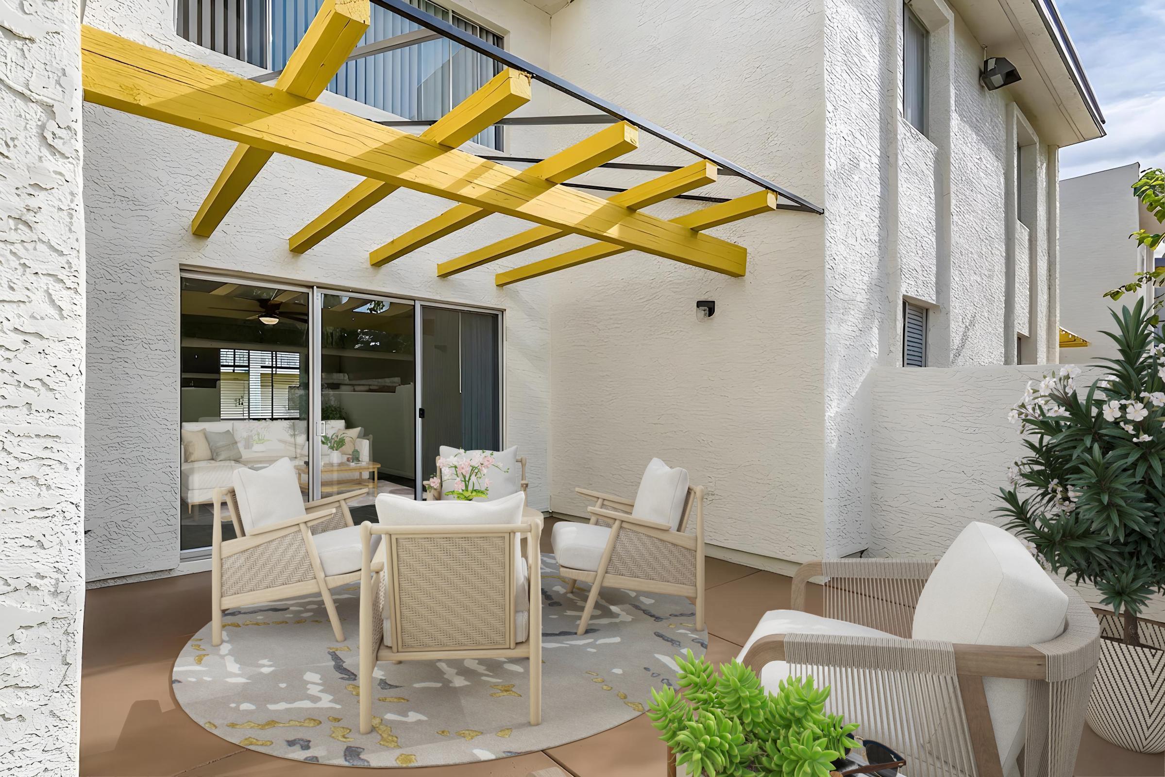 A cozy outdoor patio featuring four white chairs arranged around a round area rug. A yellow wooden pergola provides shade, and sliding glass doors lead into the interior. Potted plants and flowering bushes add greenery to the space, creating a welcoming atmosphere for relaxation.