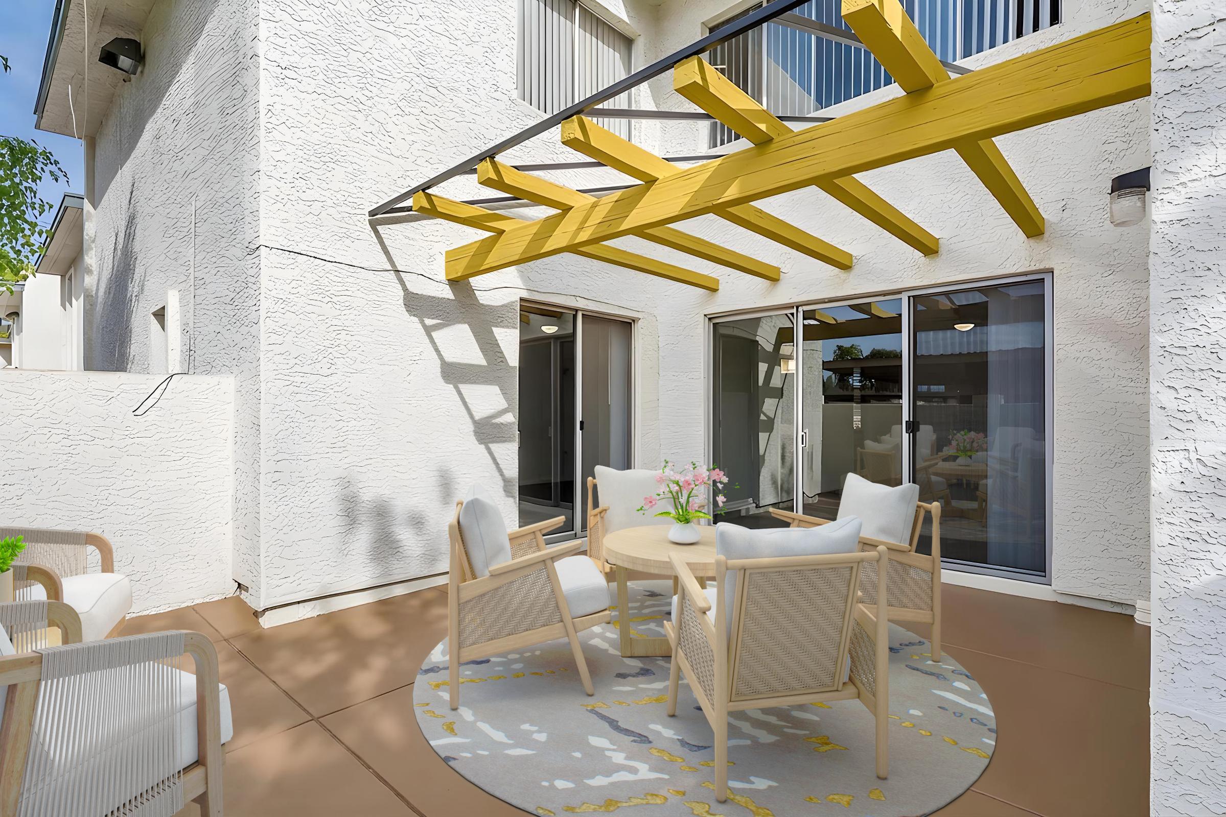 A cozy outdoor patio area featuring a round table with four beige chairs, a light-colored rug, and a bouquet of flowers. The space is surrounded by white walls and large glass doors leading into a well-lit interior, under a yellow pergola.