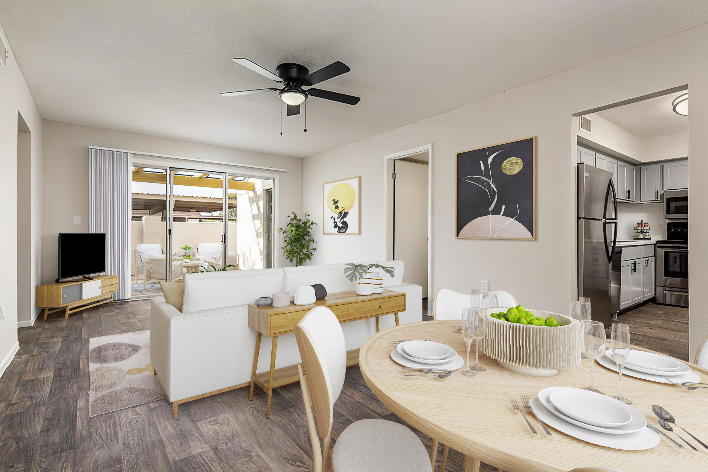 A modern living space featuring a cozy dining area with a round table set for four, white dishes, and a bowl of green apples. The adjacent living room has a white sofa, a wall-mounted TV, and decorative artwork. Large windows allow natural light, and a glimpse of an outdoor patio can be seen.