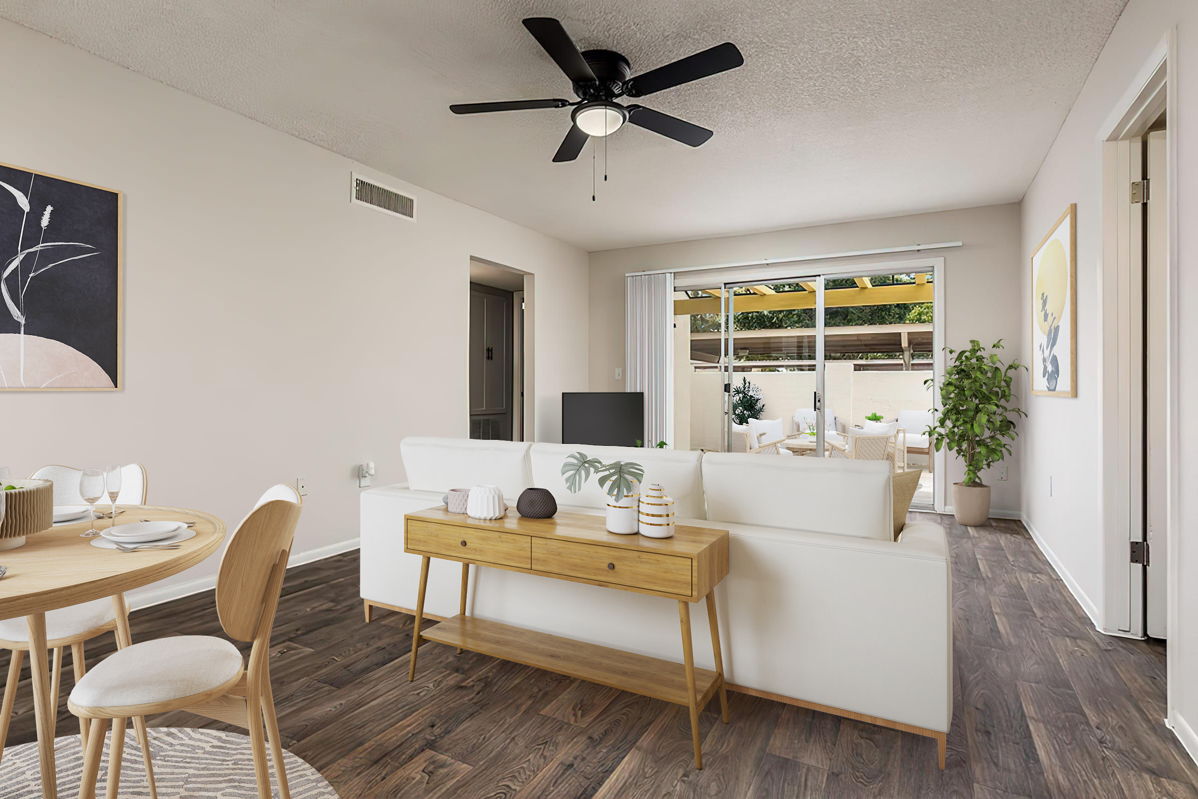 A modern living space featuring a white sofa with decorative cushions, a wooden console table with plants and decor items, a dining area with a round table and chairs, and a fan on the ceiling. Large windows allow natural light, revealing a patio area with outdoor furniture and greenery.