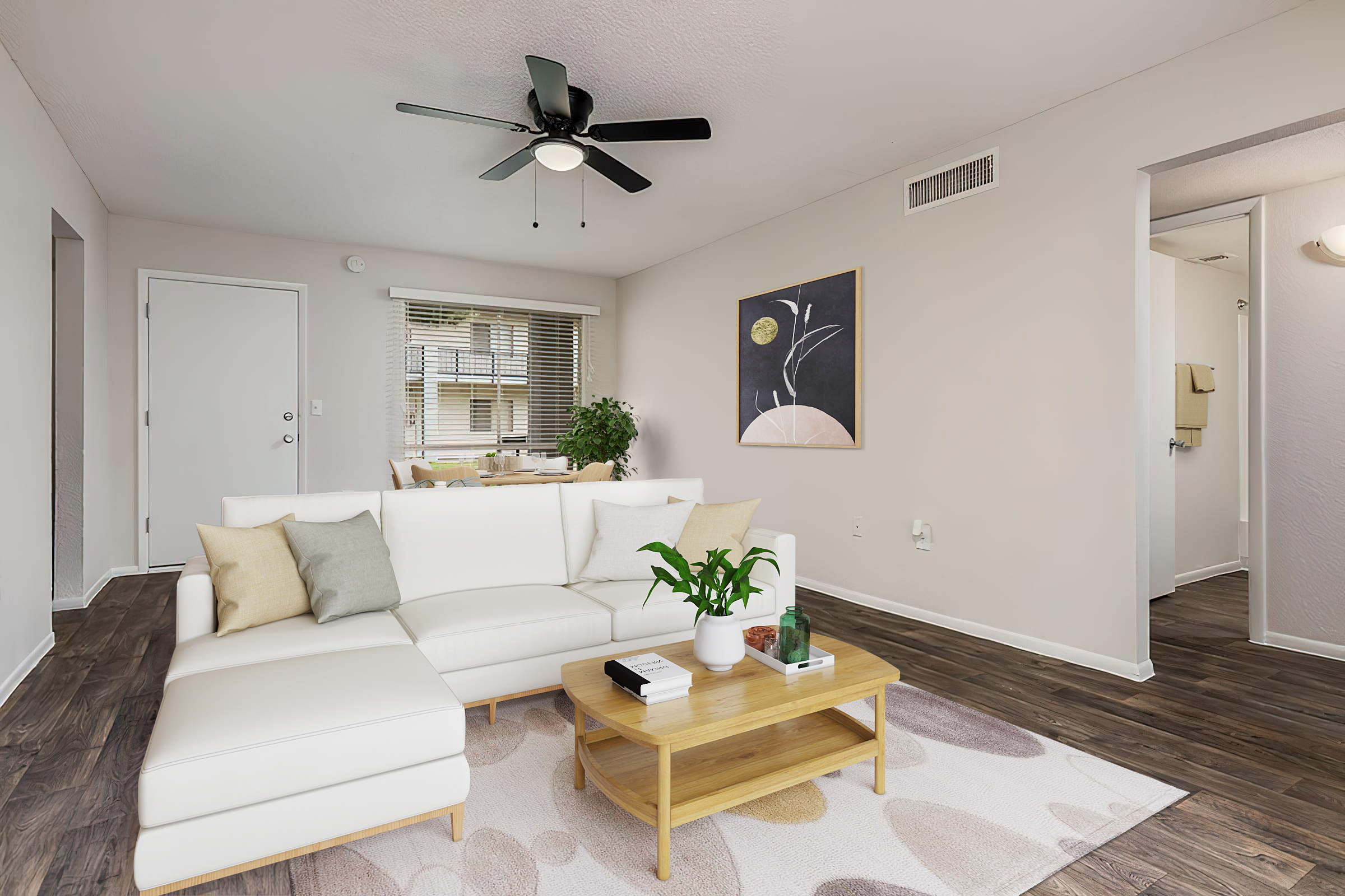 A cozy living room featuring a white sectional sofa with decorative pillows, a wooden coffee table with a plant and books, and a light rug. The walls are painted in neutral tones, and there is a ceiling fan above. A window lets in natural light, and there is artwork hanging on the wall. A doorway leads to another room.
