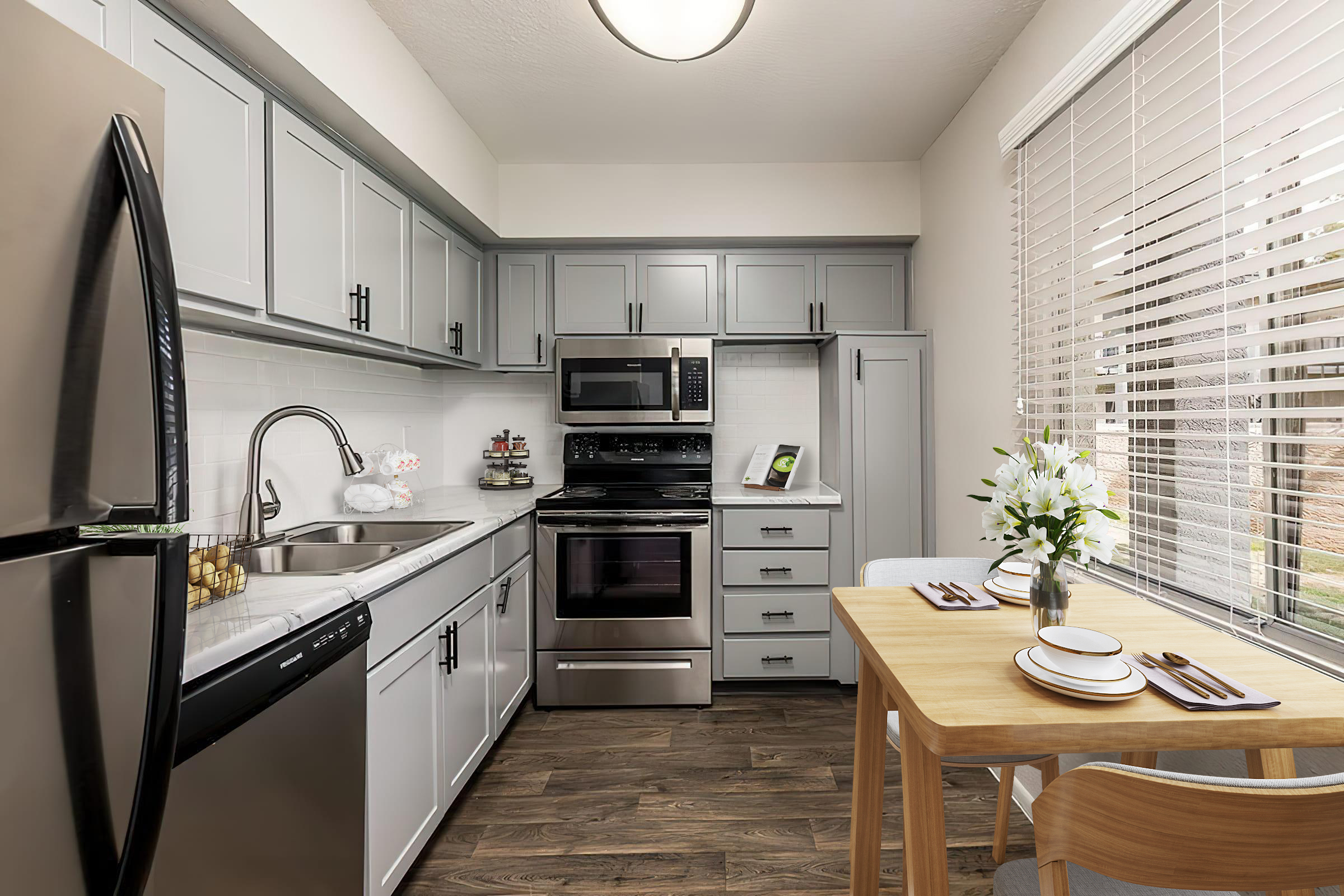 A modern kitchen featuring gray cabinetry, stainless steel appliances, and a light wood dining table. The kitchen includes a sink, oven, and microwave, with a cozy seating area near a window partially covered by blinds. Decorative elements like flowers and kitchenware enhance the space's inviting atmosphere.