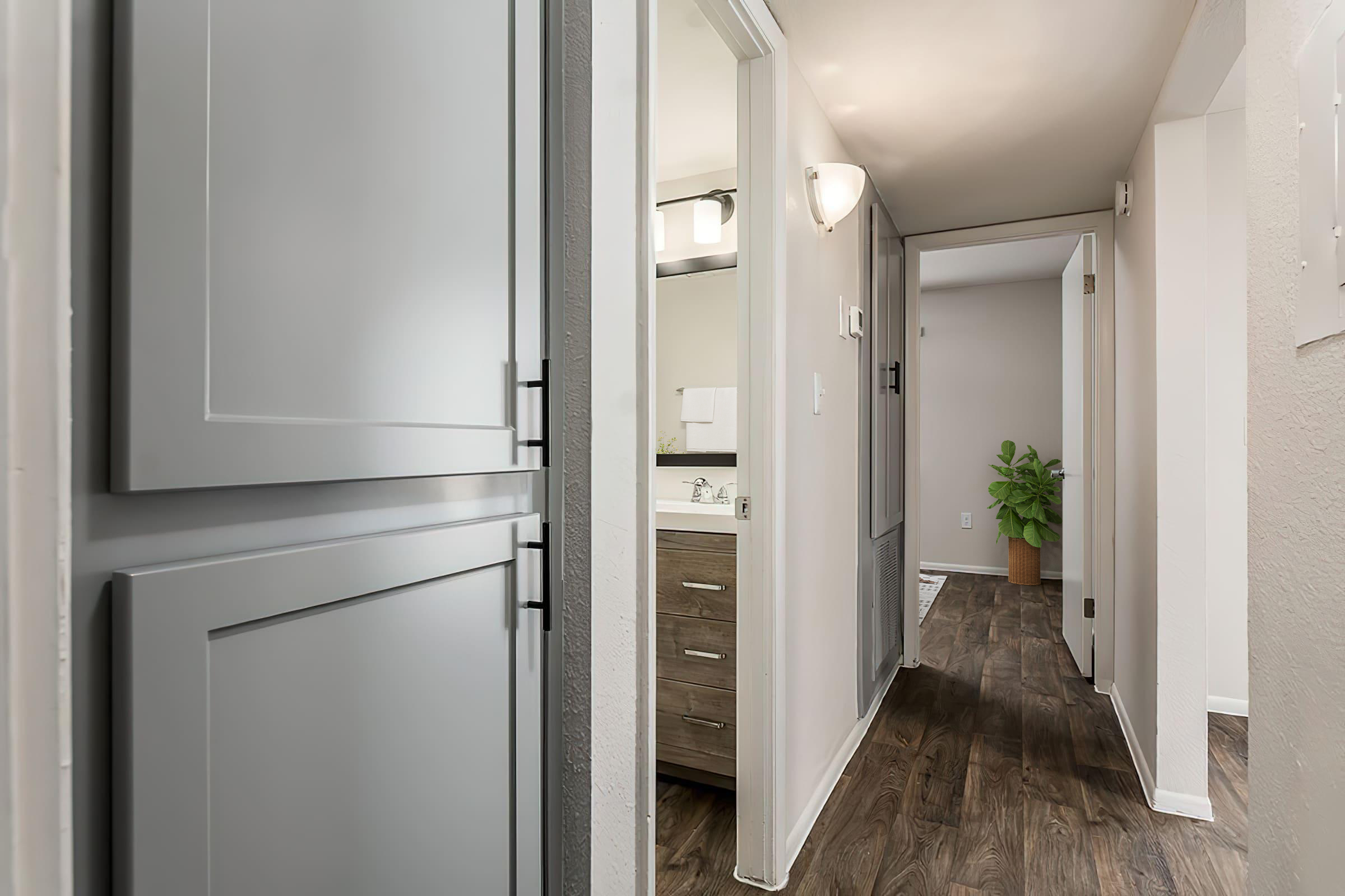 A view of a modern hallway with light-colored walls and dark wood flooring. On the left, a closed cabinet door is visible, while the end of the hallway opens to a well-lit bathroom with a mirror and vanity. A potted plant adds a touch of greenery to the scene.