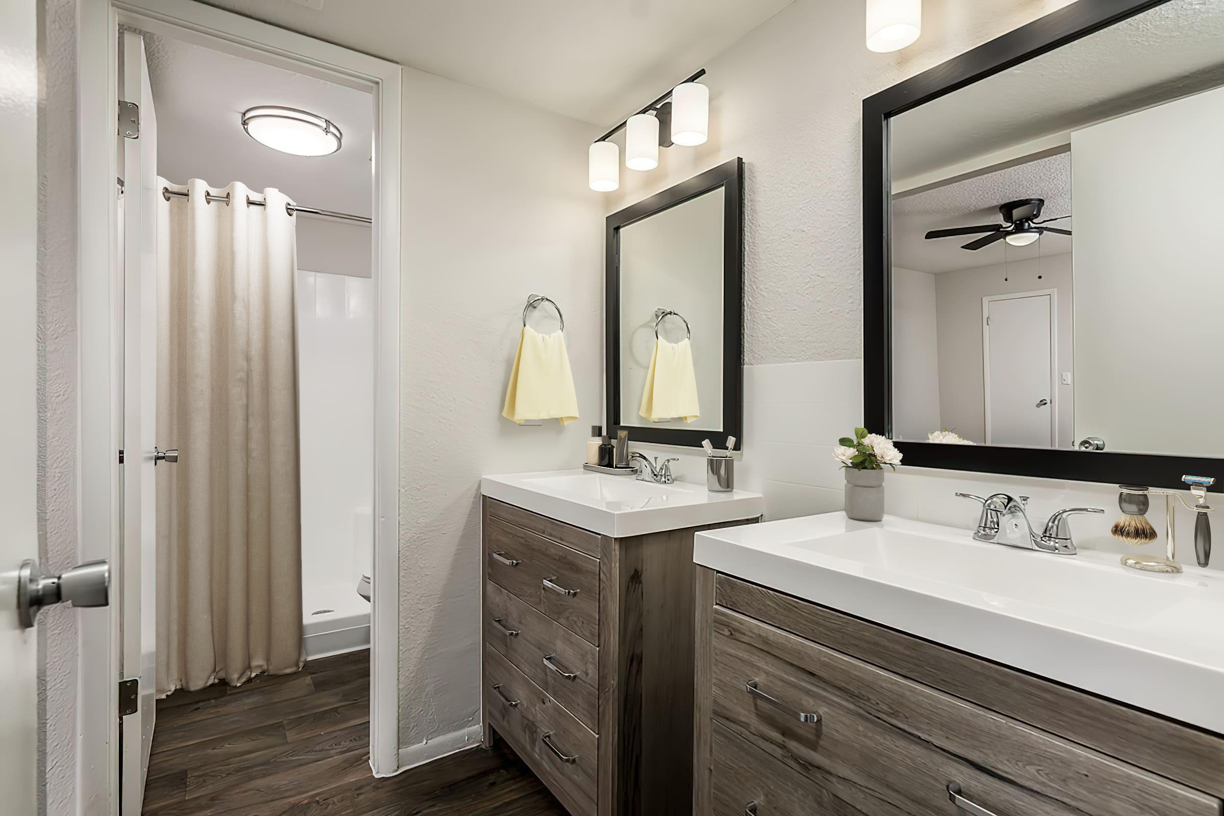A modern bathroom featuring a double sink vanity with wooden cabinets and two mirrors. There is a shower area with a beige curtain, a light fixture with multiple bulbs, and a towel rack with yellow towels. The wall colors are neutral, and the flooring is dark wood.