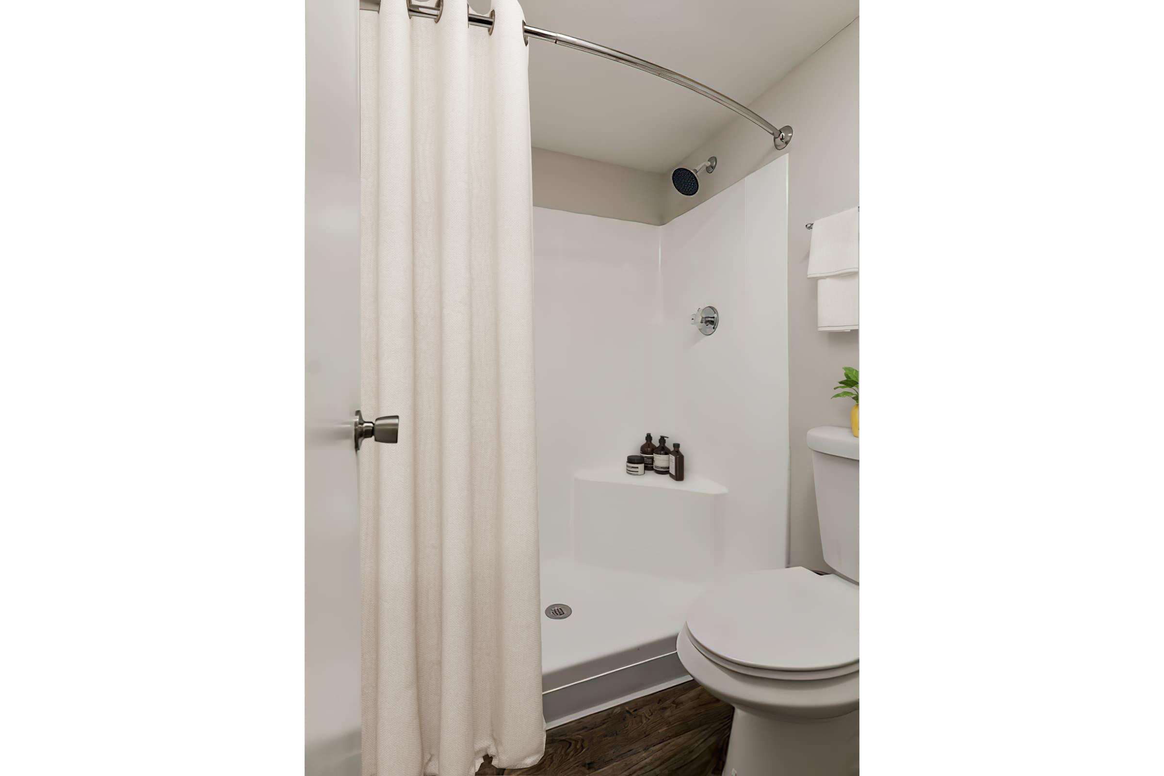 A clean, modern bathroom with a shower featuring a curved curtain rod and a white shower curtain. The shower area has a built-in shelf with toiletries, and there's a toilet beside it. Light neutral colors create a bright and spacious feel.