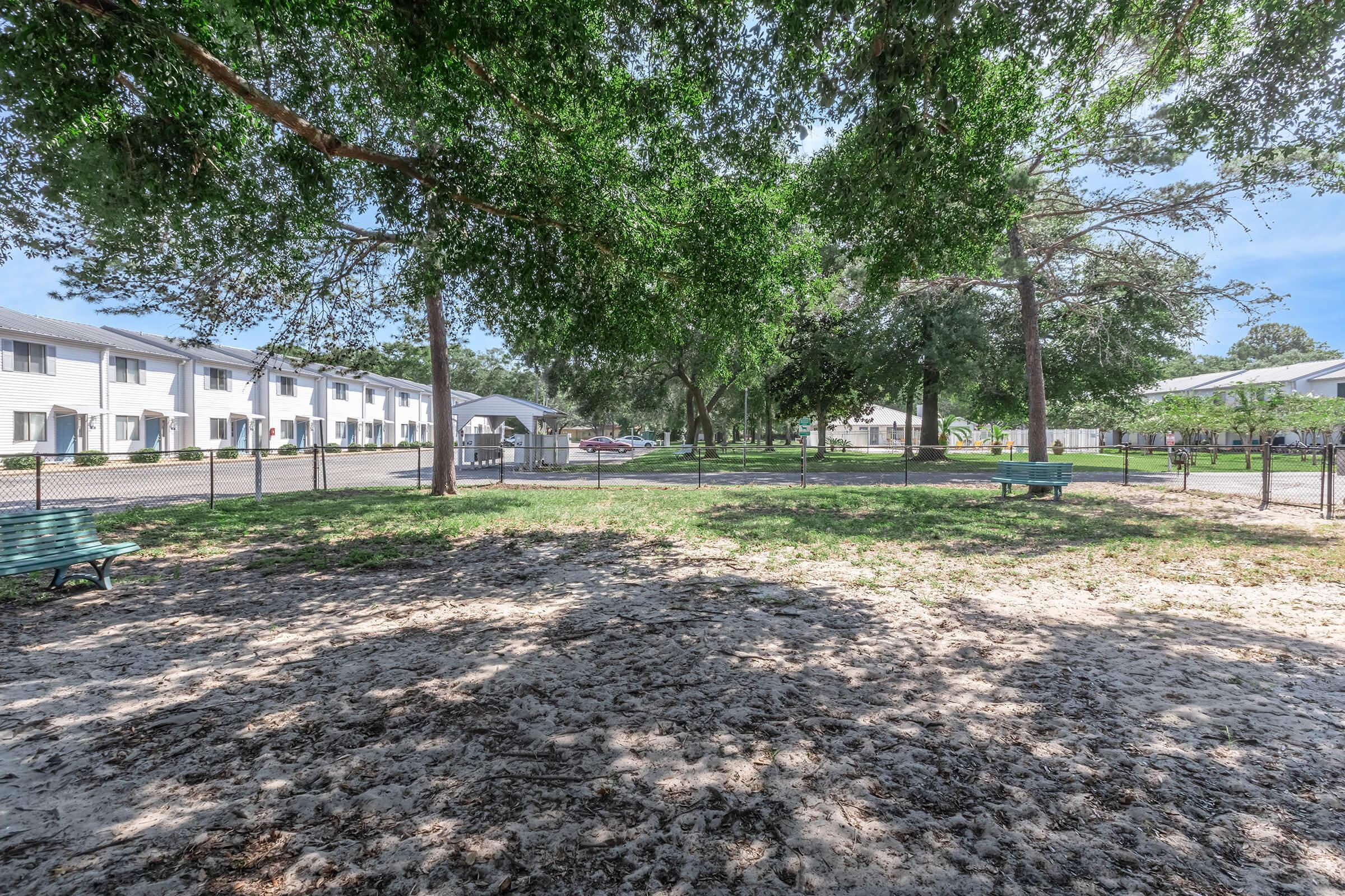 A spacious park area featuring benches, sandy ground, and shade from large trees. In the background, a row of apartment buildings and a parking area are visible. The scene is bright and inviting, suggesting a communal outdoor space for relaxation and socializing.