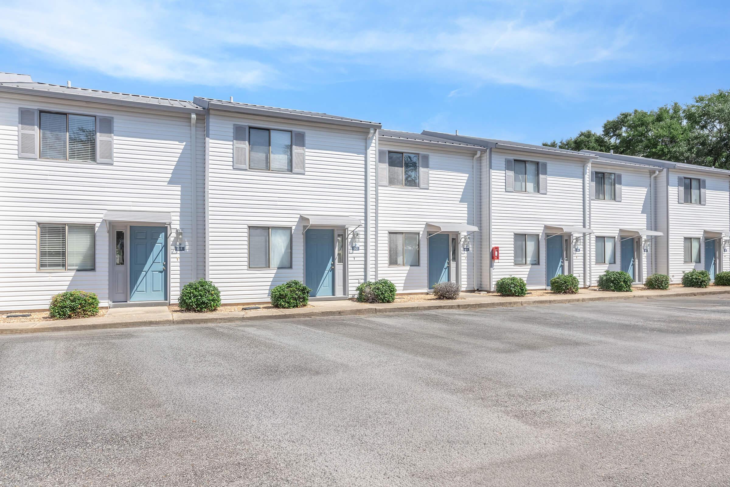 A row of modern, two-story townhouses with light-colored siding and blue doors. The street is paved and lined with small shrubs. Clear blue sky in the background, creating a bright and inviting atmosphere.