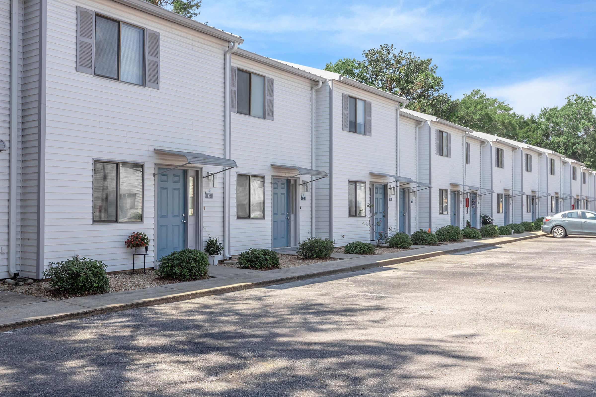 Row of white, two-story townhouses with blue doors and windows. Each unit has a small porch area with potted plants, and the parking lot in front features a few parked cars. The scene is landscaped with greenery and trees in the background under a clear blue sky.