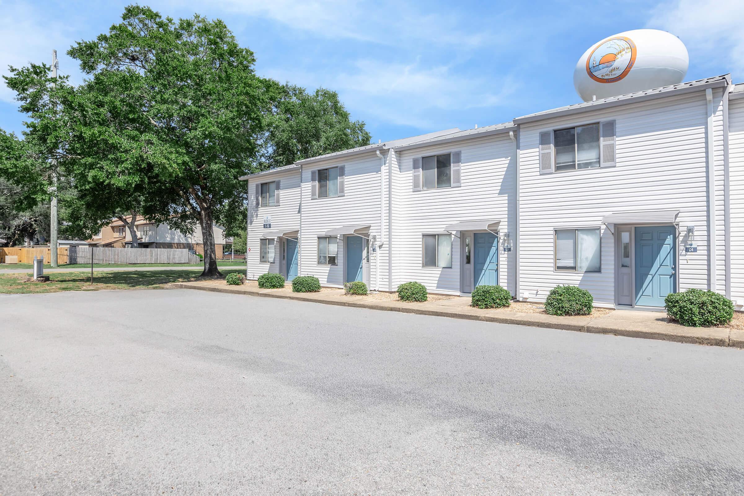 Row of townhouses with light-colored siding, blue front doors, and neatly trimmed bushes in front. A large water tower with a circular design is visible in the background. A paved driveway leads up to the buildings under a bright blue sky.