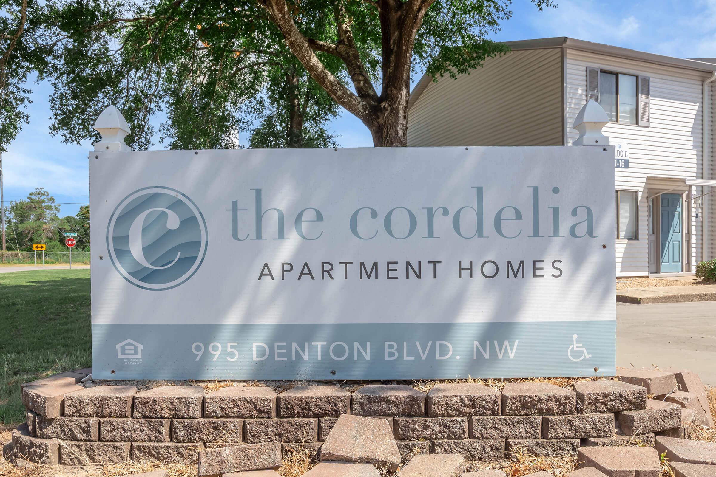 Sign for The Cordelia Apartment Homes, located at 995 Denton Blvd. NW. The sign features a logo representing the apartment complex, with trees and a grassy area in the background. It indicates accessibility options.