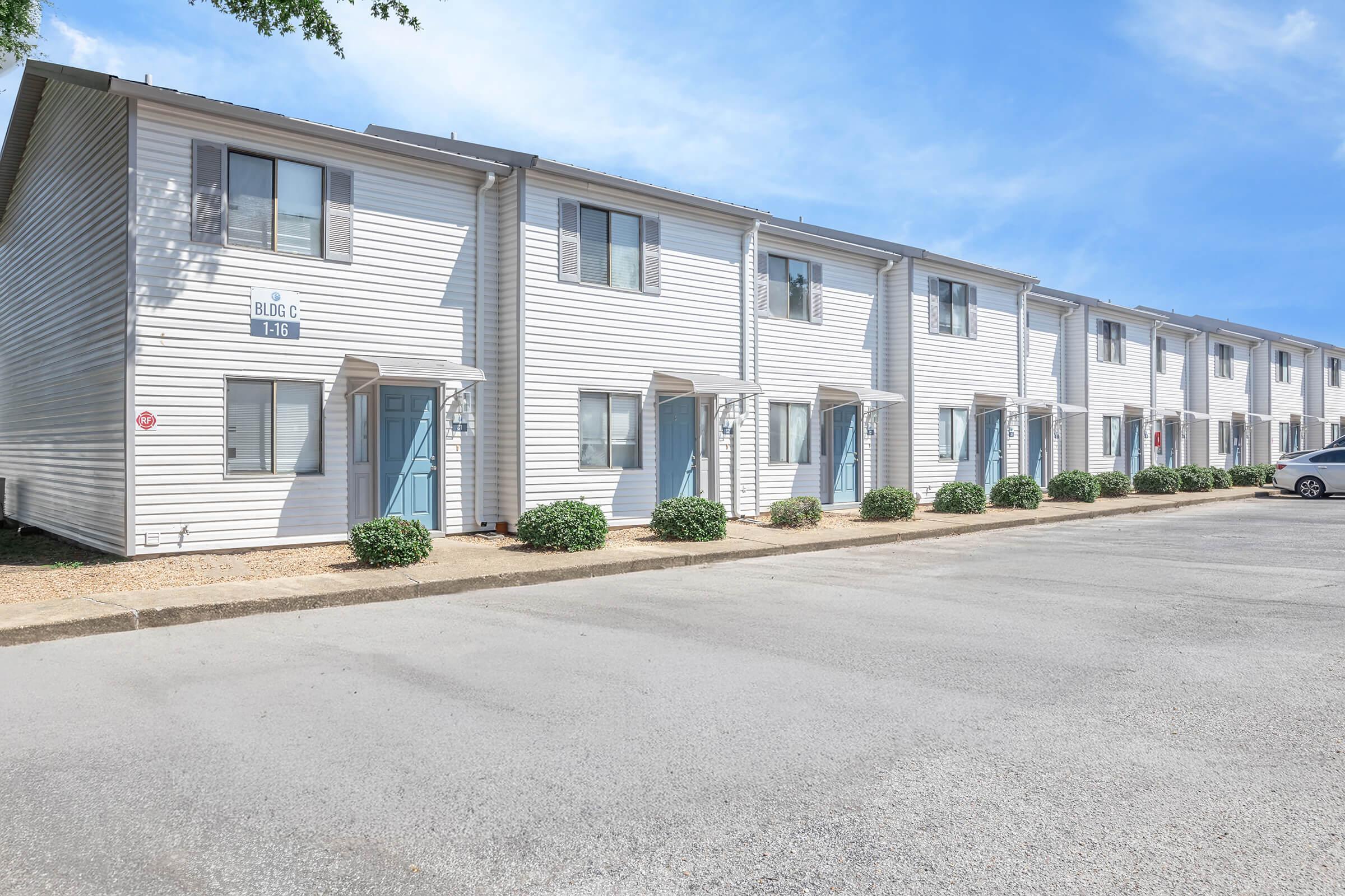 A row of white two-story apartment buildings with blue doors and small shrubs in front. The parking area is visible, and the sky is bright and clear.