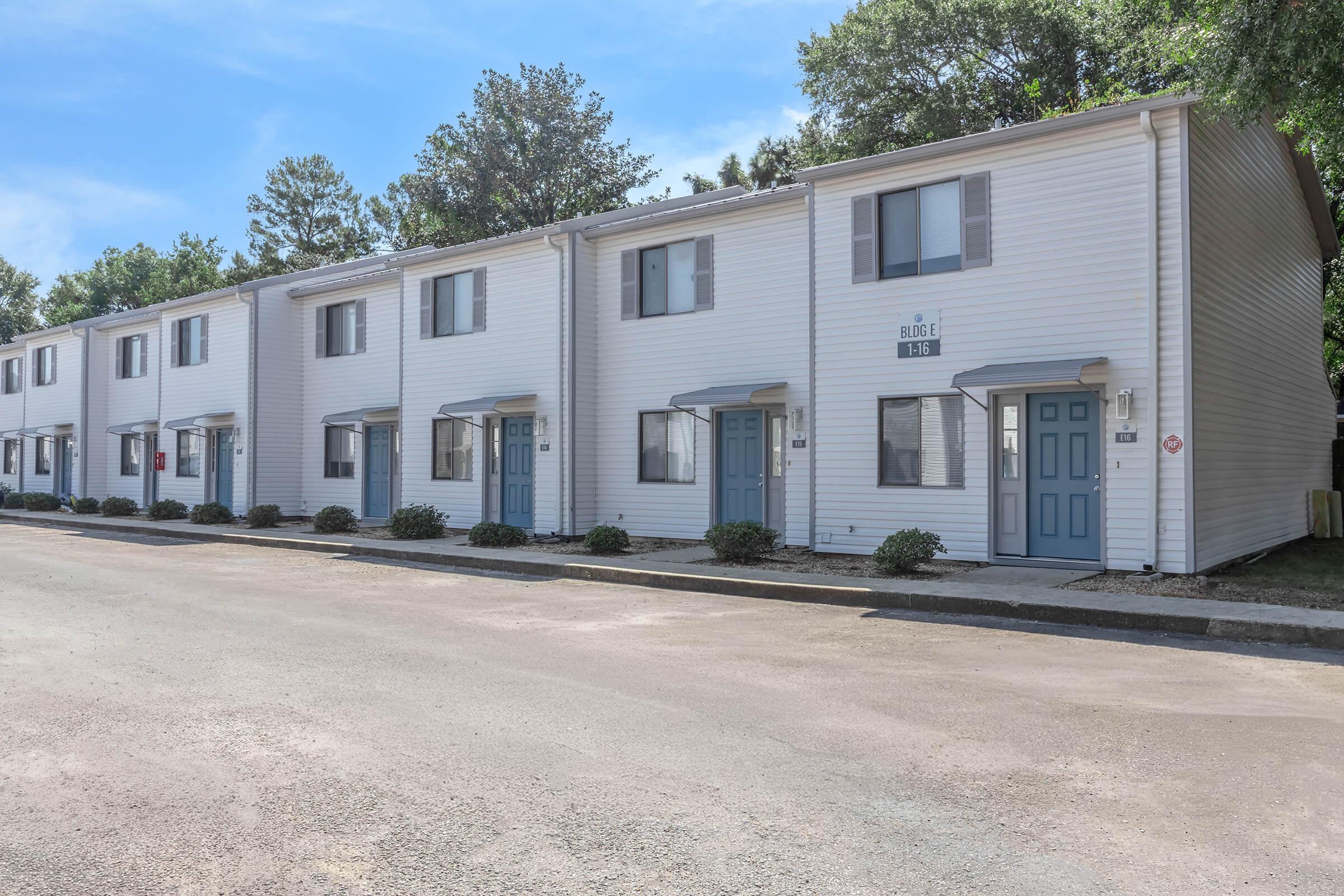 Row of two-story residential buildings with white siding and blue doors, lined along a paved road. Green shrubs are planted in front of each unit, and trees are visible in the background under a clear blue sky. The scene conveys a clean, well-maintained housing area.