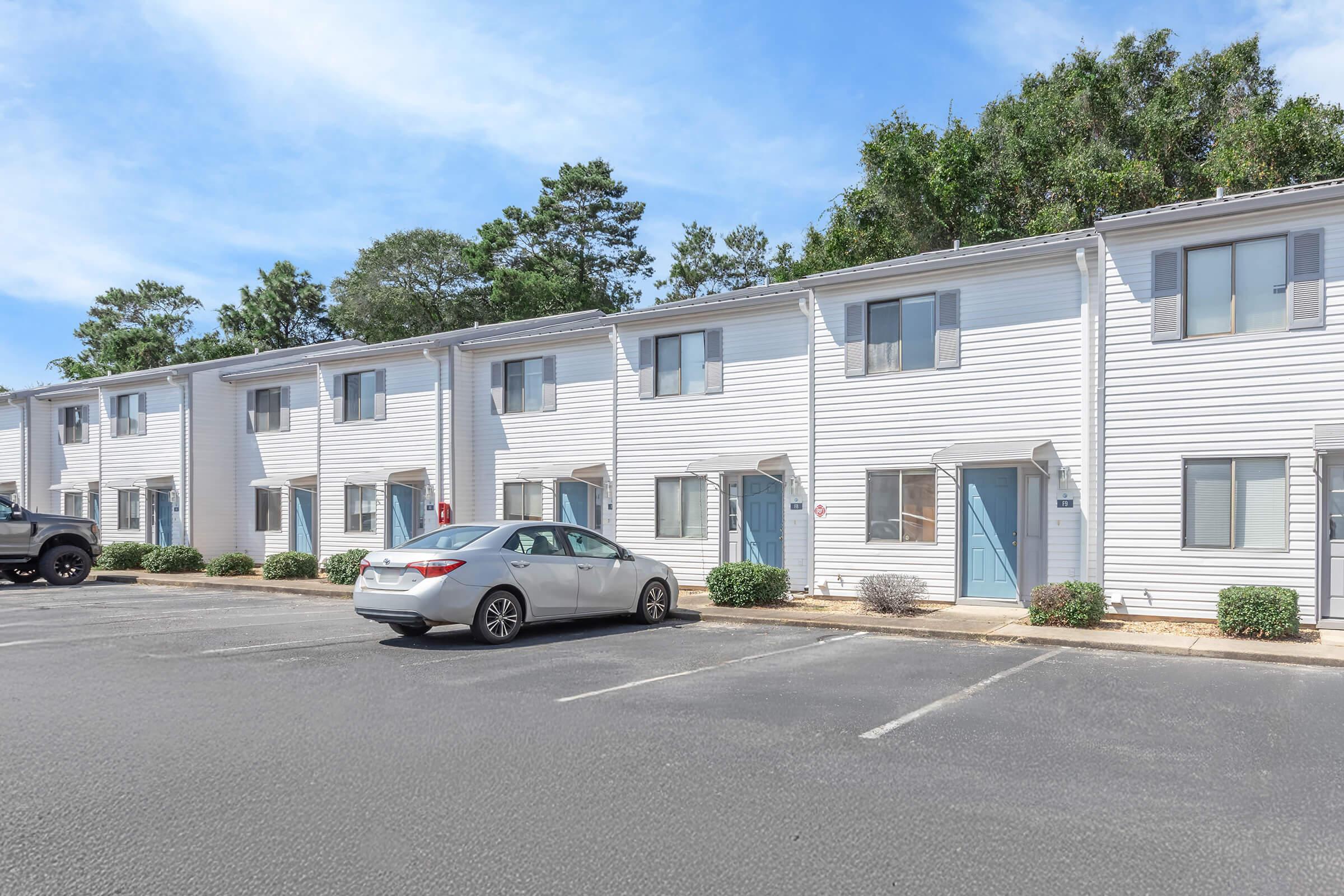 A row of white apartment buildings with blue doors, situated in a parking lot. A parked silver car is visible in front of the buildings. Lush green trees provide a backdrop, under a clear blue sky. The setting appears well maintained and inviting.