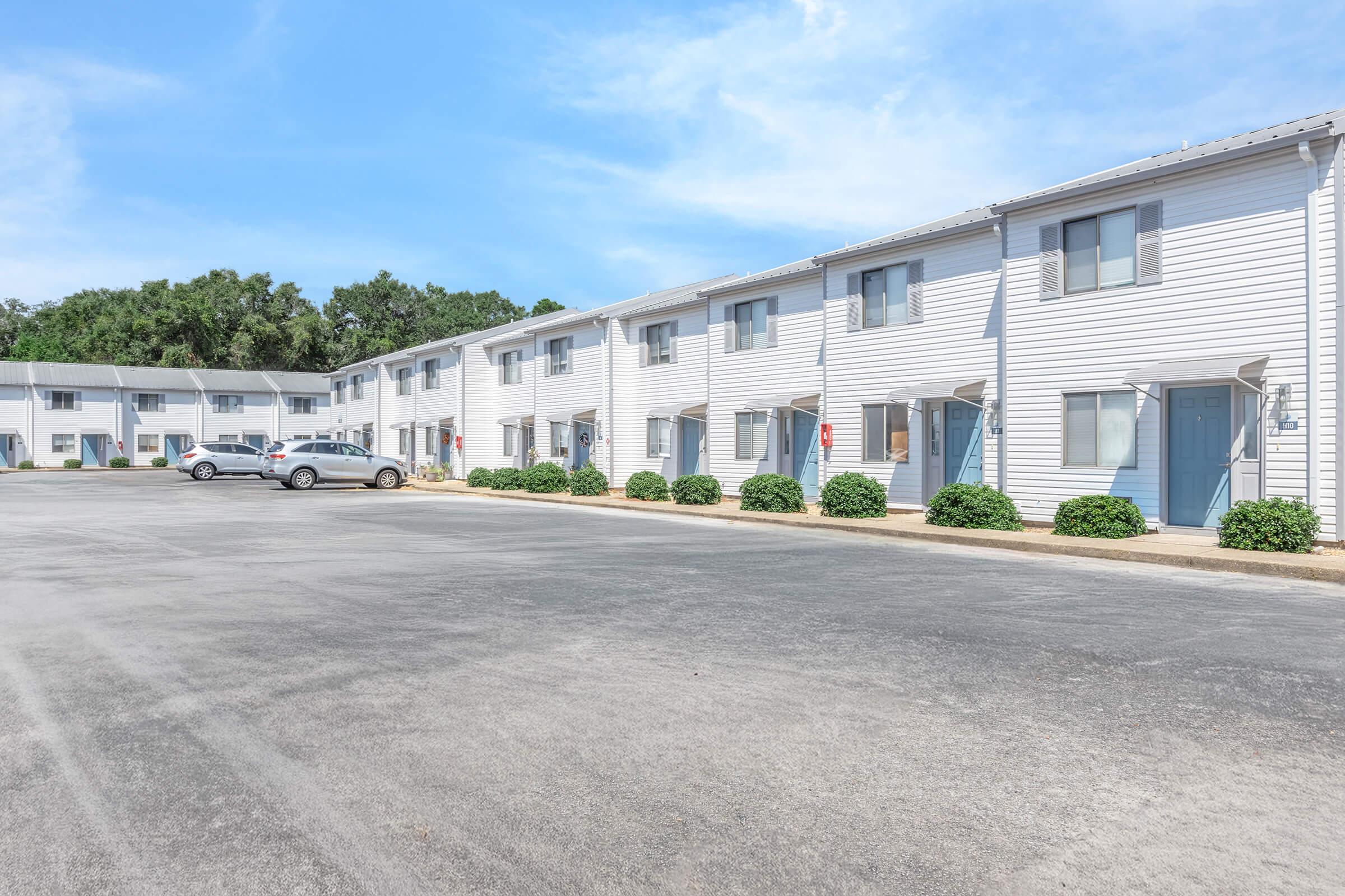 A row of two-story apartment buildings with white siding, featuring blue doors. The foreground shows a paved parking area with several cars parked. Neatly trimmed bushes line the walkway, and a clear blue sky is visible in the background, indicating a sunny day.