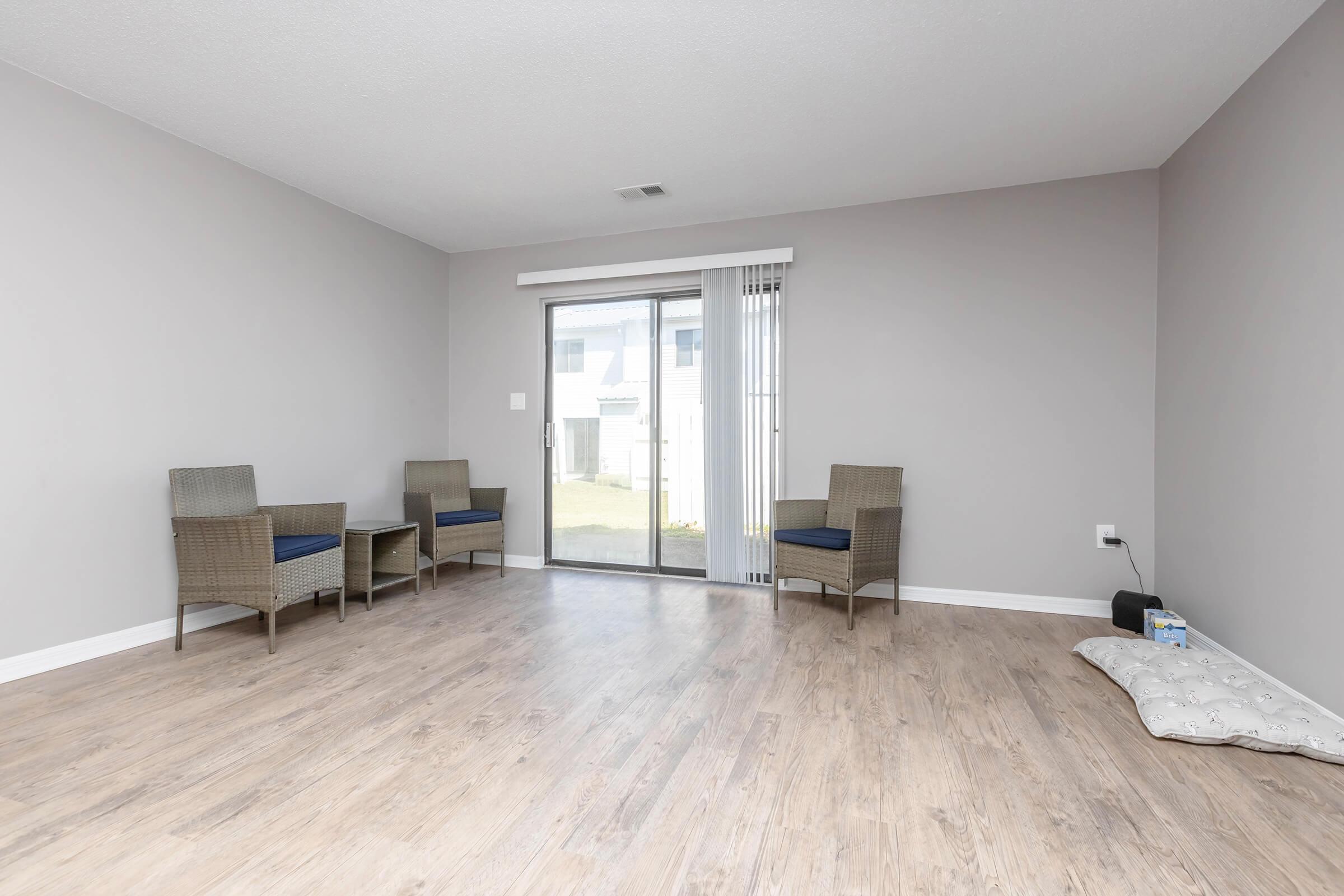 Empty living room with light gray walls, wooden flooring, and two wicker chairs positioned side by side. A sliding glass door leads to an outdoor area, with a small white pet bed on the floor. Natural light illuminates the space, creating a bright and airy atmosphere.