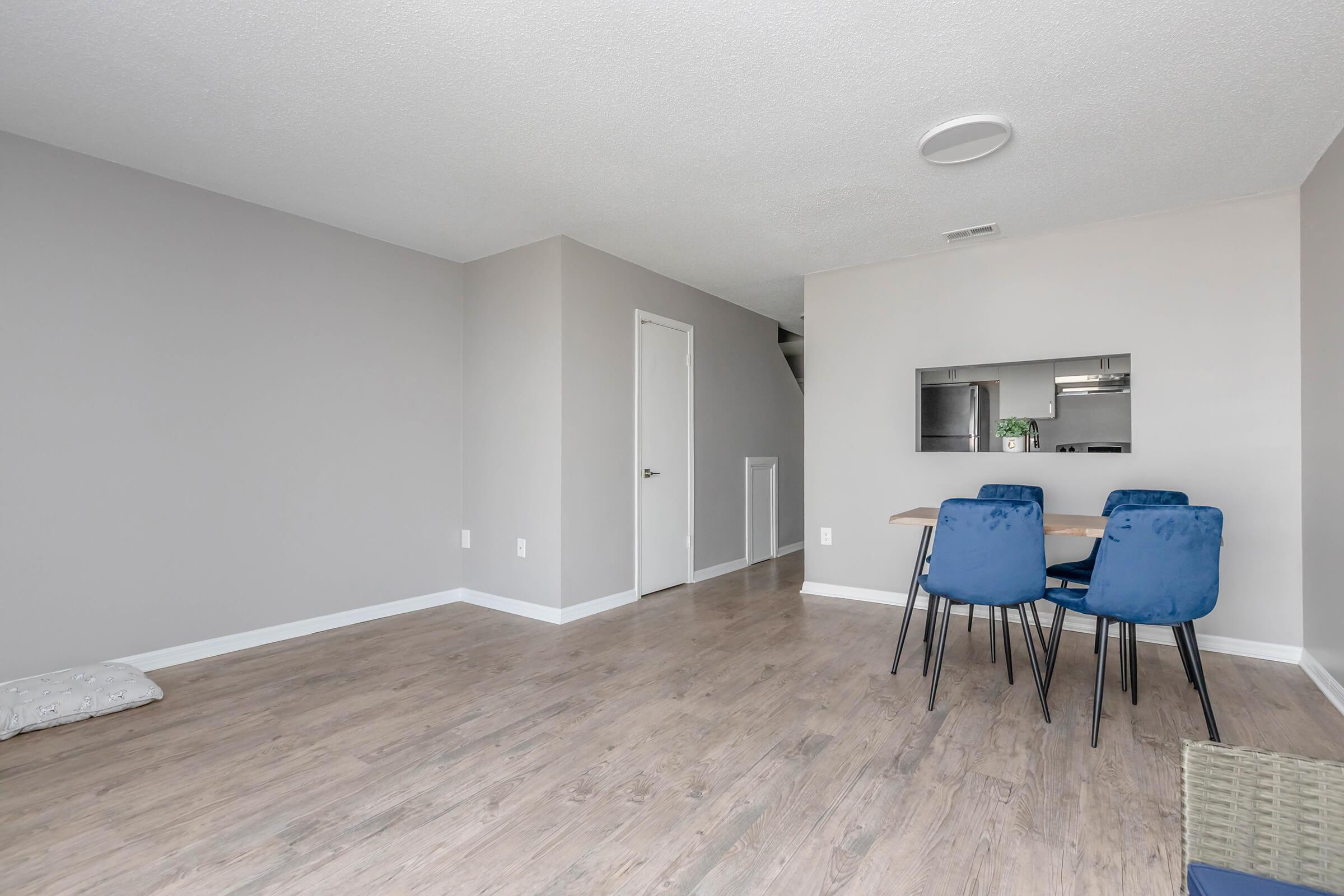 A modern, minimalistic living space featuring light gray walls and wooden flooring. A small dining area with a table and four blue chairs is visible. In the background, there is a glimpse of a kitchen, and a closed white door is seen on the left. A cozy pet bed is placed on the floor in the foreground.