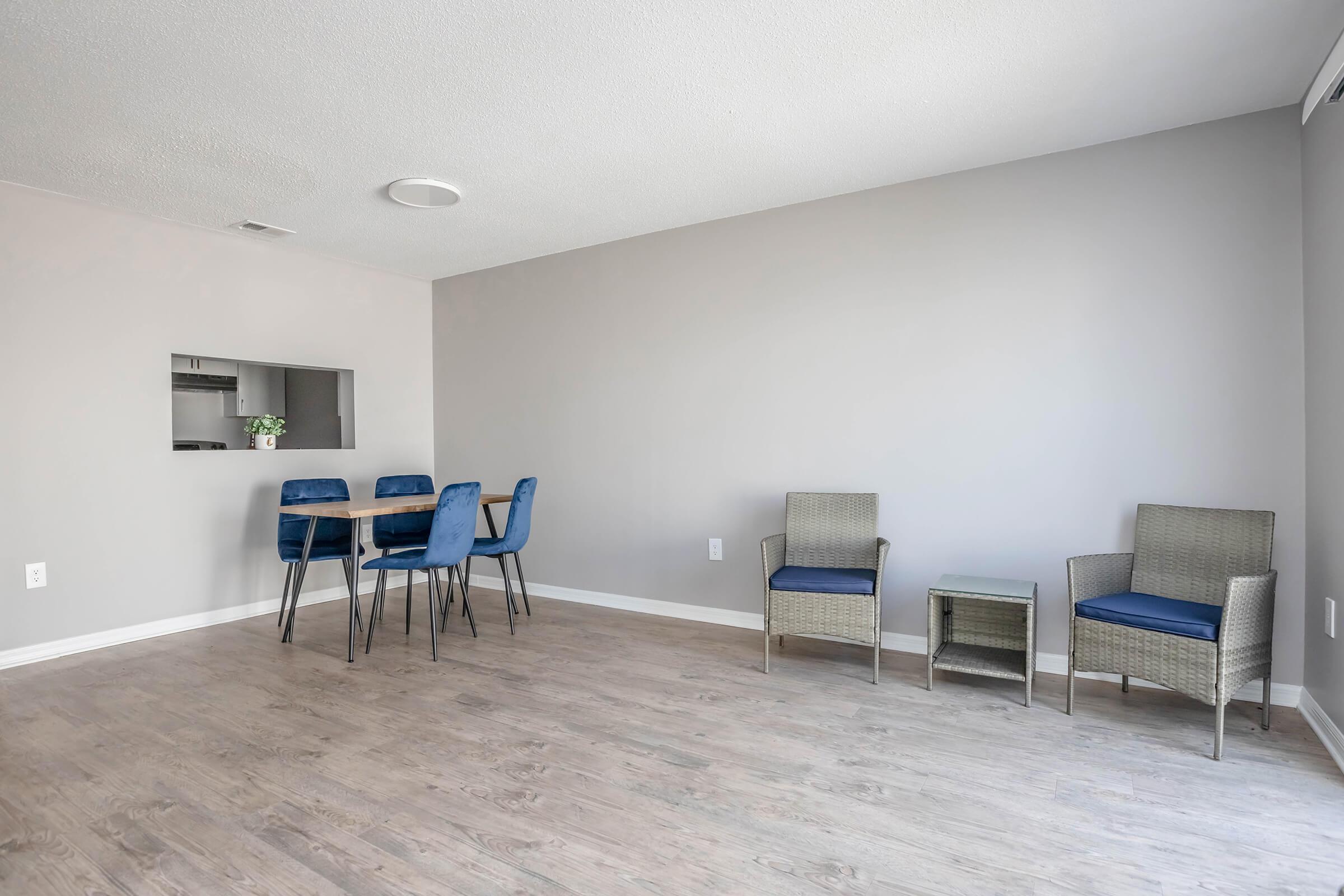 A modern living space featuring light wood flooring, a light gray painted wall, and a small dining area with a wooden table and blue chairs. To the right, there are two comfortable chairs and a small side table. A kitchenette is visible in the background through an open space in the wall.